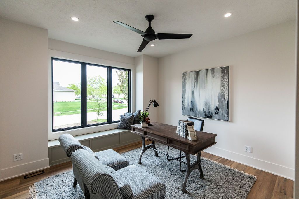 Modern office features a dark wood desk with iron legs, a gray upholstered lounge chair and ottoman, and a built-in window seat. The room has a neutral color palette with a large abstract painting on the wall and a black ceiling fan.