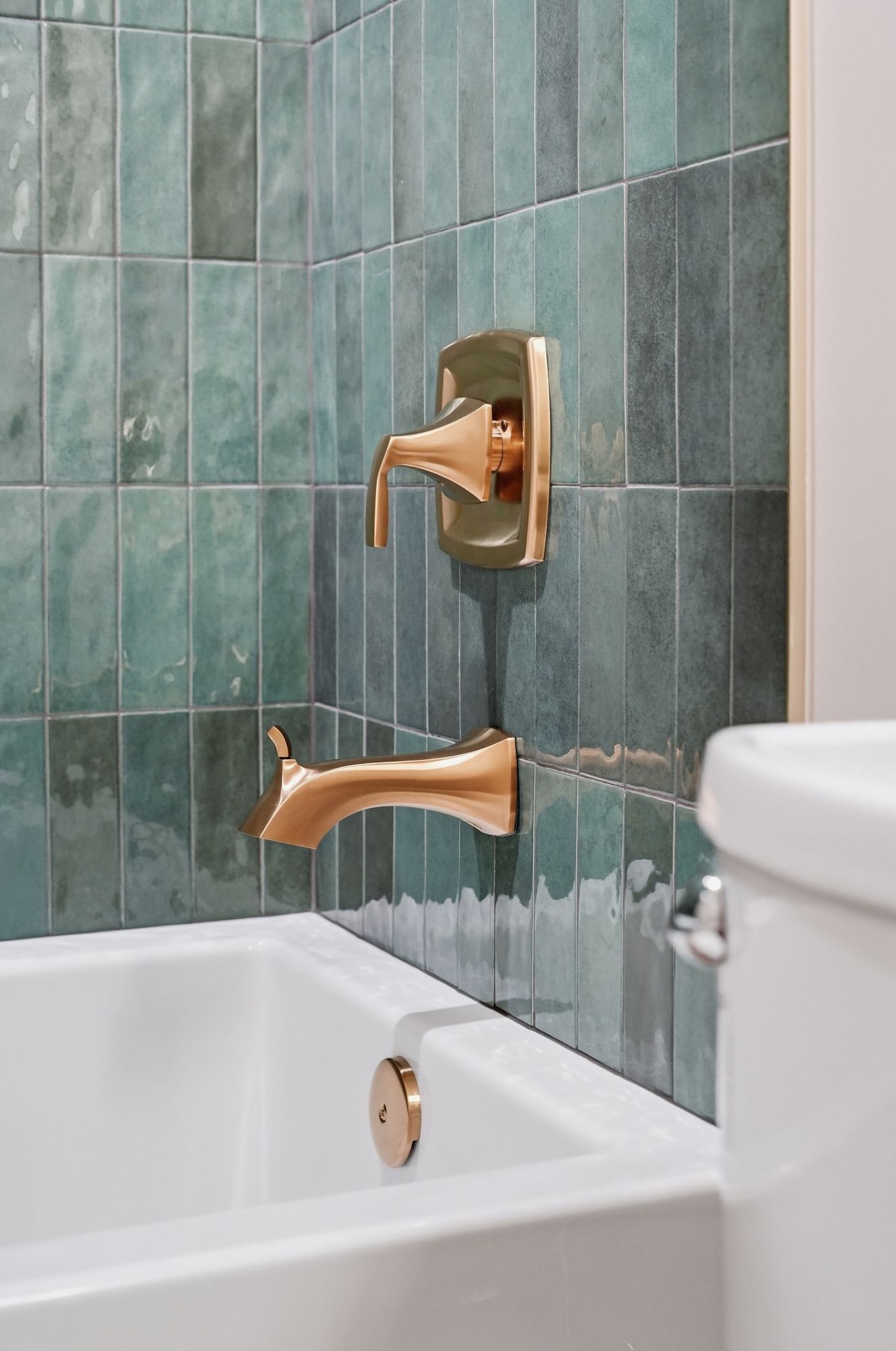 Close-up of a bathtub with gold fixtures, including a faucet and drain plug, against a green tiled wall. A portion of a white toilet is visible on the right side of the frame.