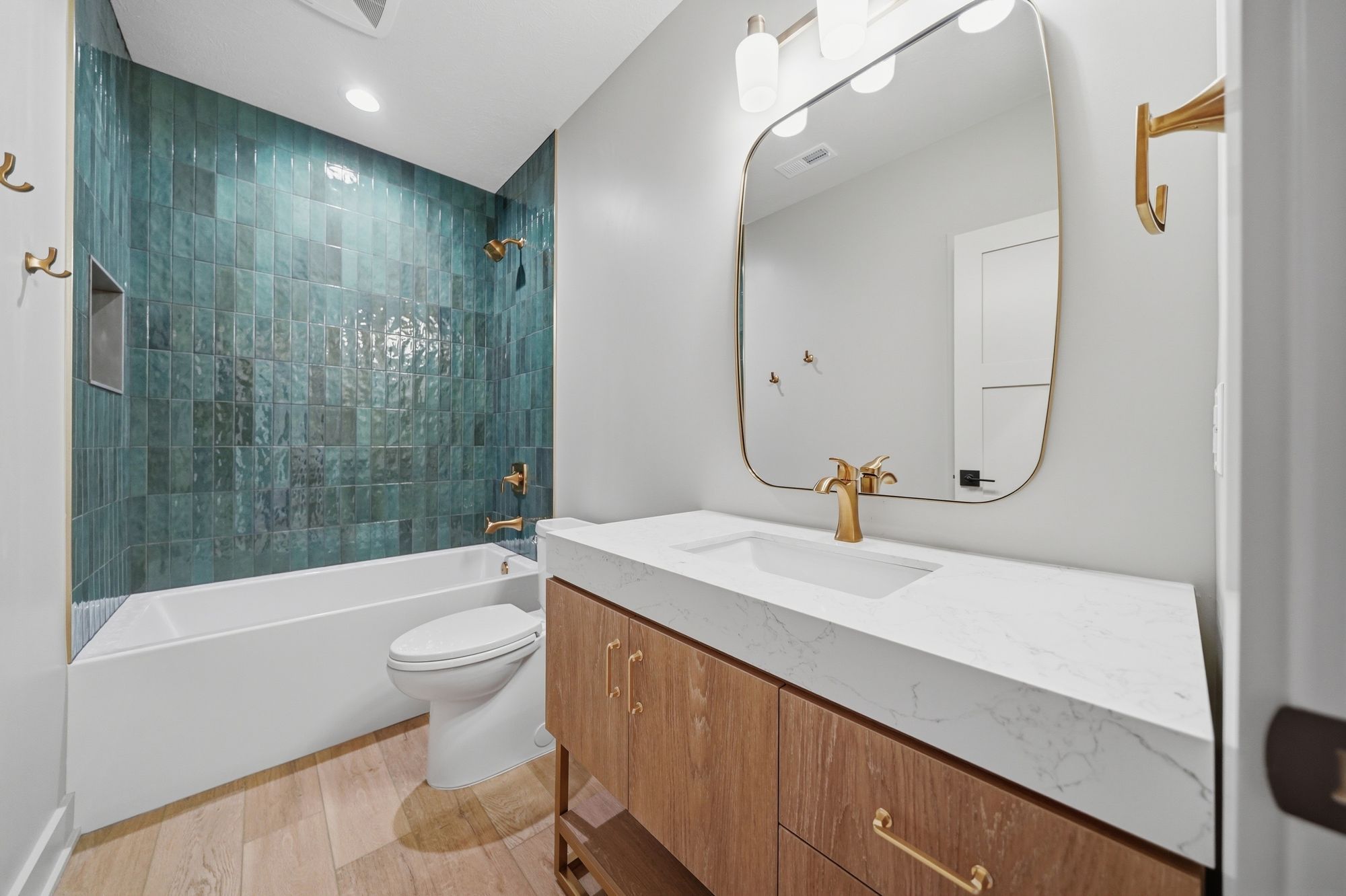 Modern bathroom with wood-look flooring and striking teal tiled shower surround. The vanity features a white countertop, wood-grain cabinetry, and gold fixtures, complementing the gold-framed mirror and matching hardware.
