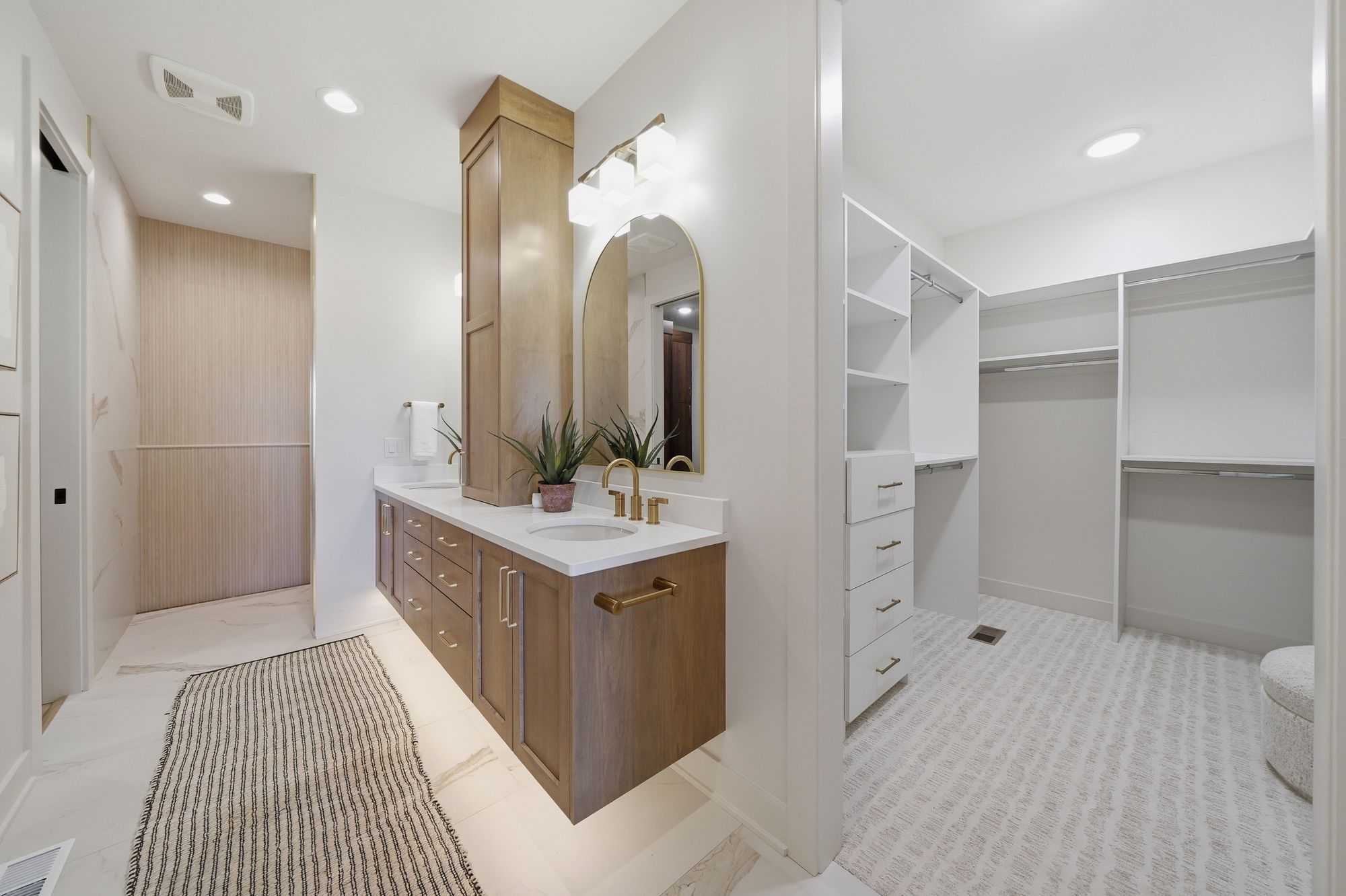 Bright bathroom with wood vanity and gold fixtures adjoins a walk-in closet featuring white shelving units and carpet. The vanity features a white countertop, gold faucet, and an arched mirror with a gold frame.
