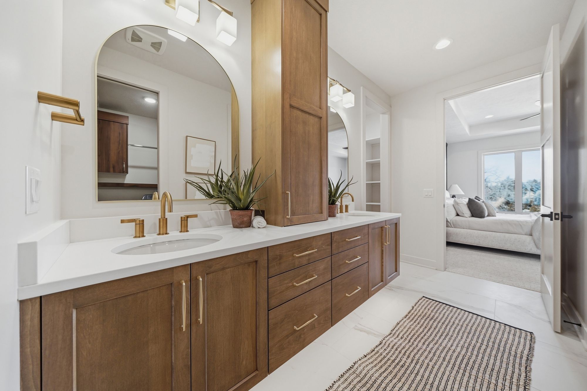 Bright bathroom featuring dual sinks with white countertops and light brown wood cabinetry, arched mirrors with gold frames, and gold hardware. The open doorway reveals a bedroom with a bed, window, and soft natural light.