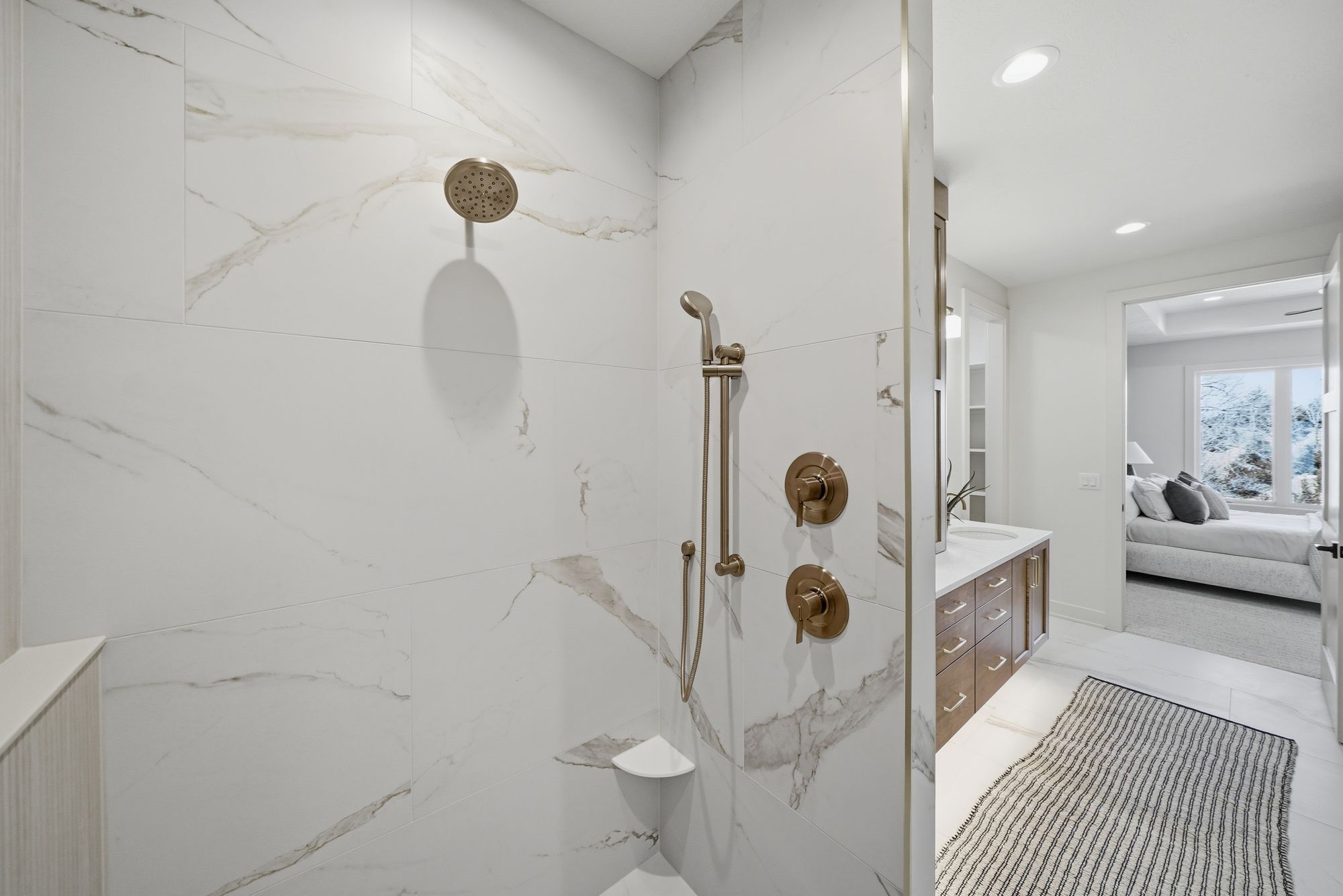 Modern bathroom features a shower with marble-look tile, brushed gold fixtures, and a built-in soap dish. A striped rug leads to a vanity area and a bedroom with a wintery view.