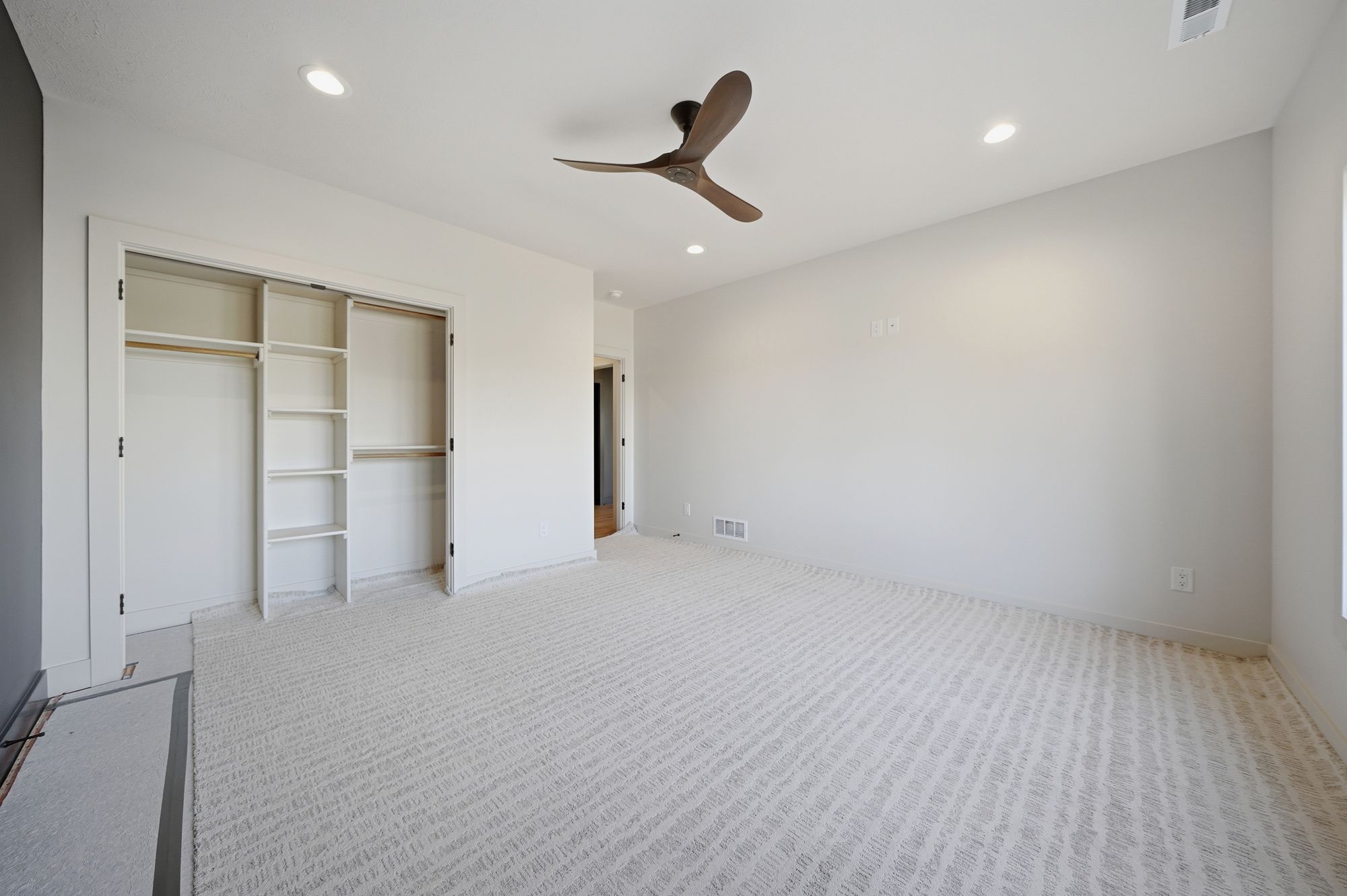 Bright bedroom with neutral decor includes a ceiling fan, closet, and patterned carpet. The room has white walls and multiple recessed lights.