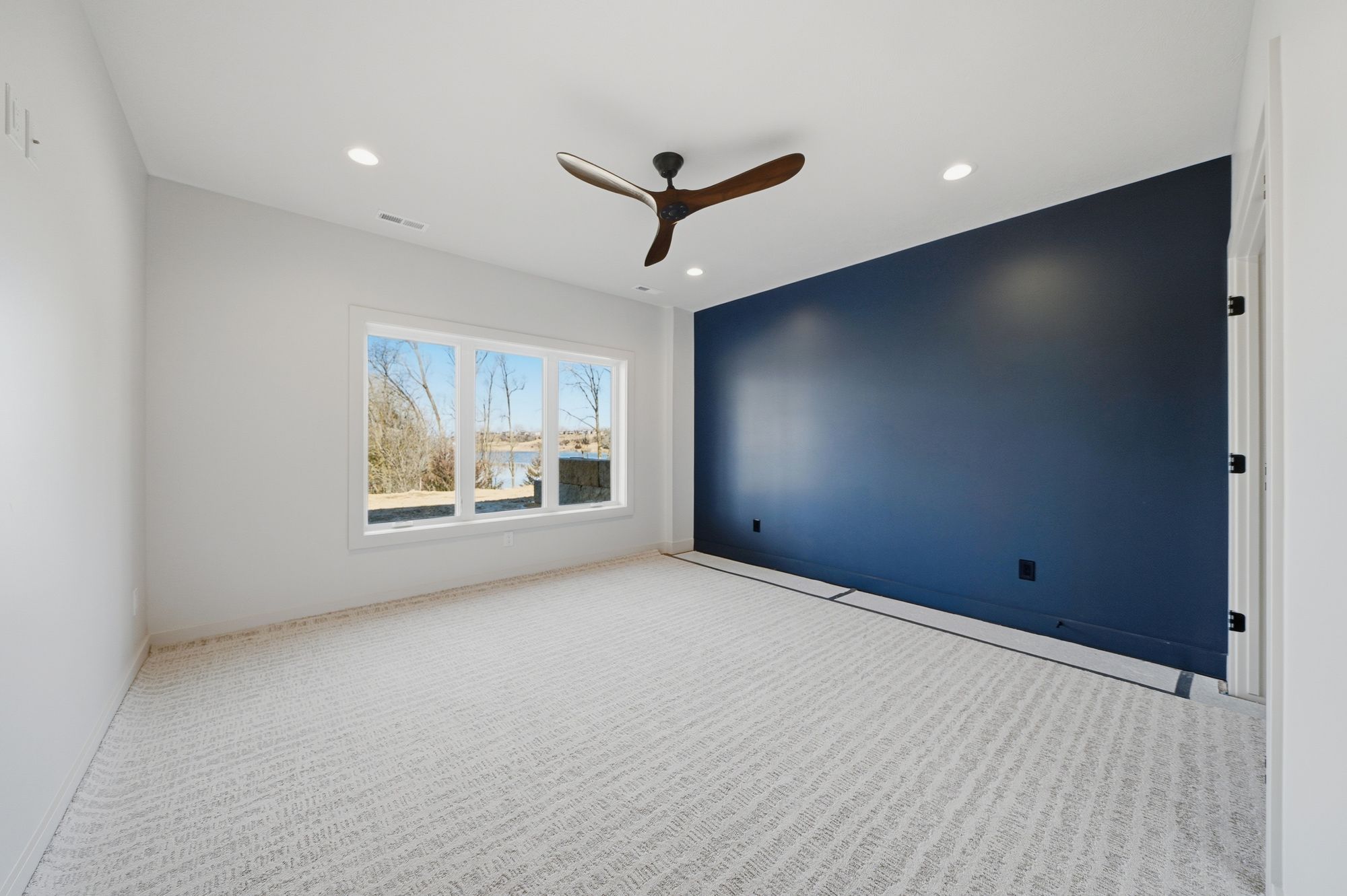 Modern bedroom with a large window providing natural light and a statement navy blue accent wall. The room features neutral carpet and a unique ceiling fan.