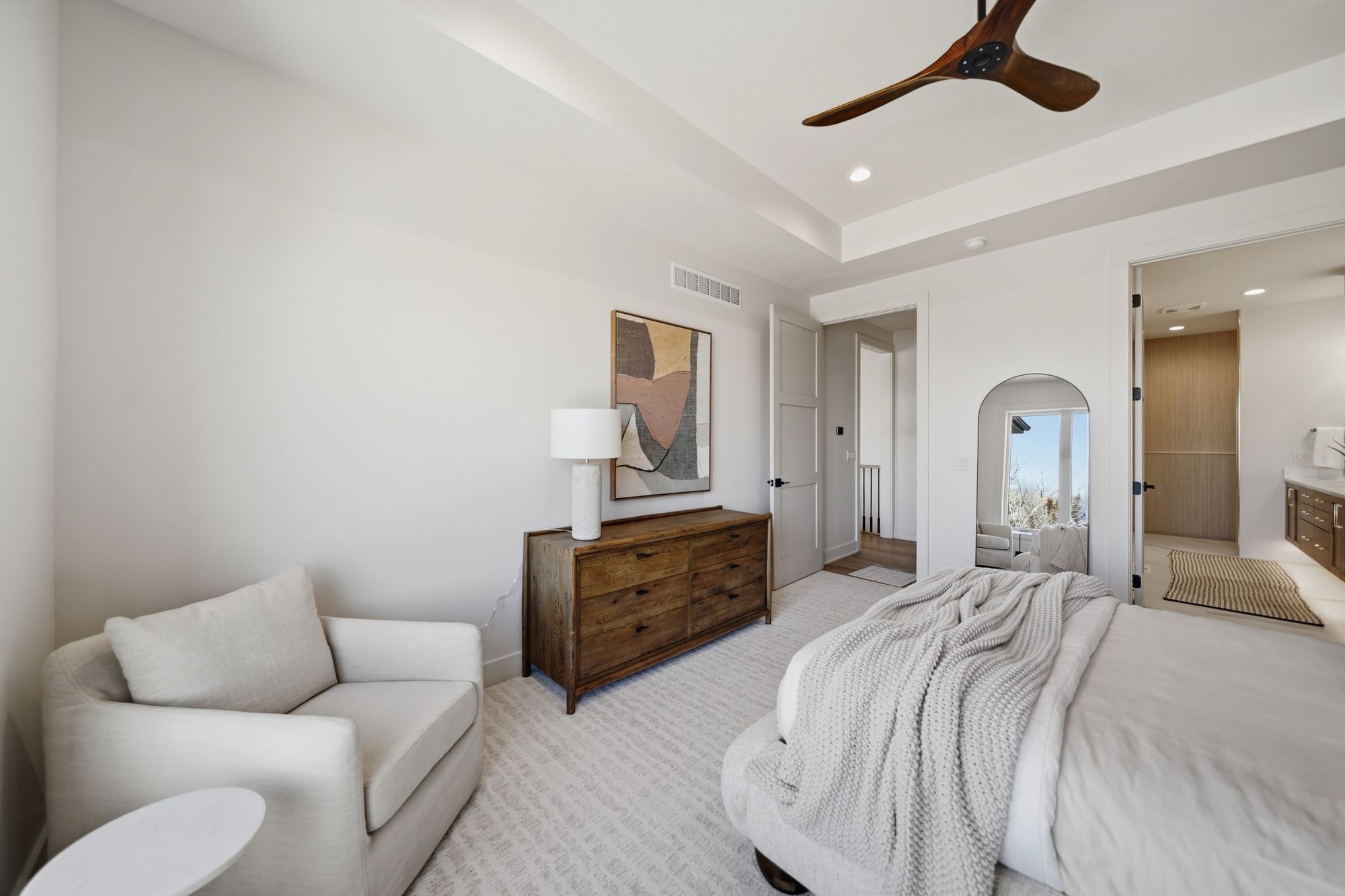 This bedroom features a neutral palette with a modern aesthetic. It includes a wooden dresser, an abstract painting, a comfortable armchair, and a large bed with a cozy blanket.