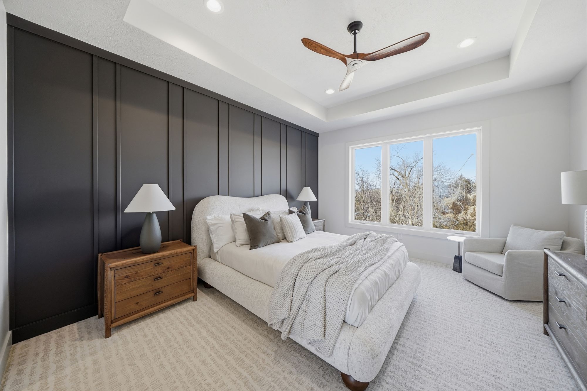 This bedroom features a statement dark-gray paneled wall, a neutral upholstered bed, and minimalist decor. A ceiling fan and recessed lighting illuminate the space, complemented by natural light from a large window with a scenic view.