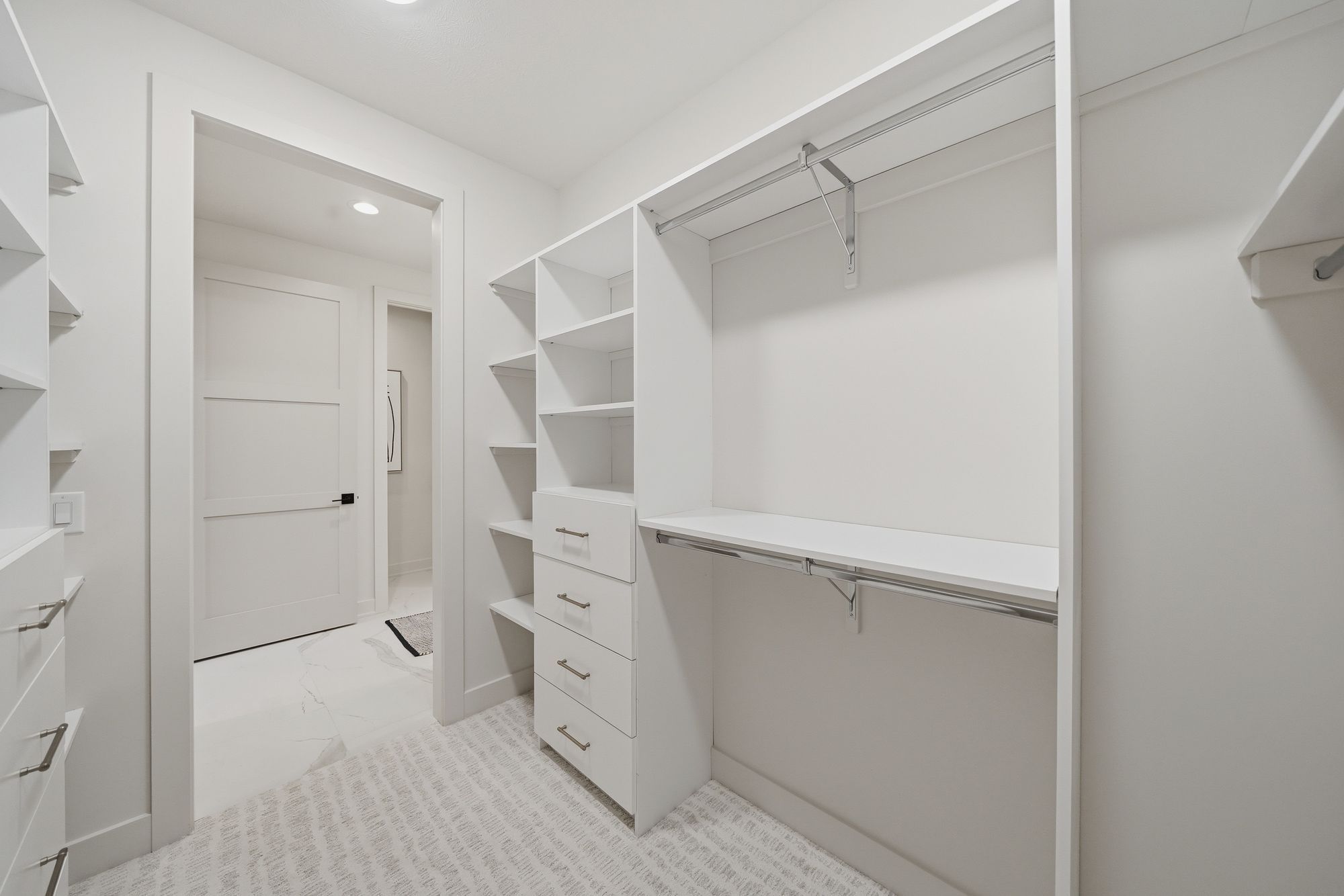Bright walk-in closet with custom shelving and drawers in an all-white color scheme. A doorway leads to another room with white marble flooring.
