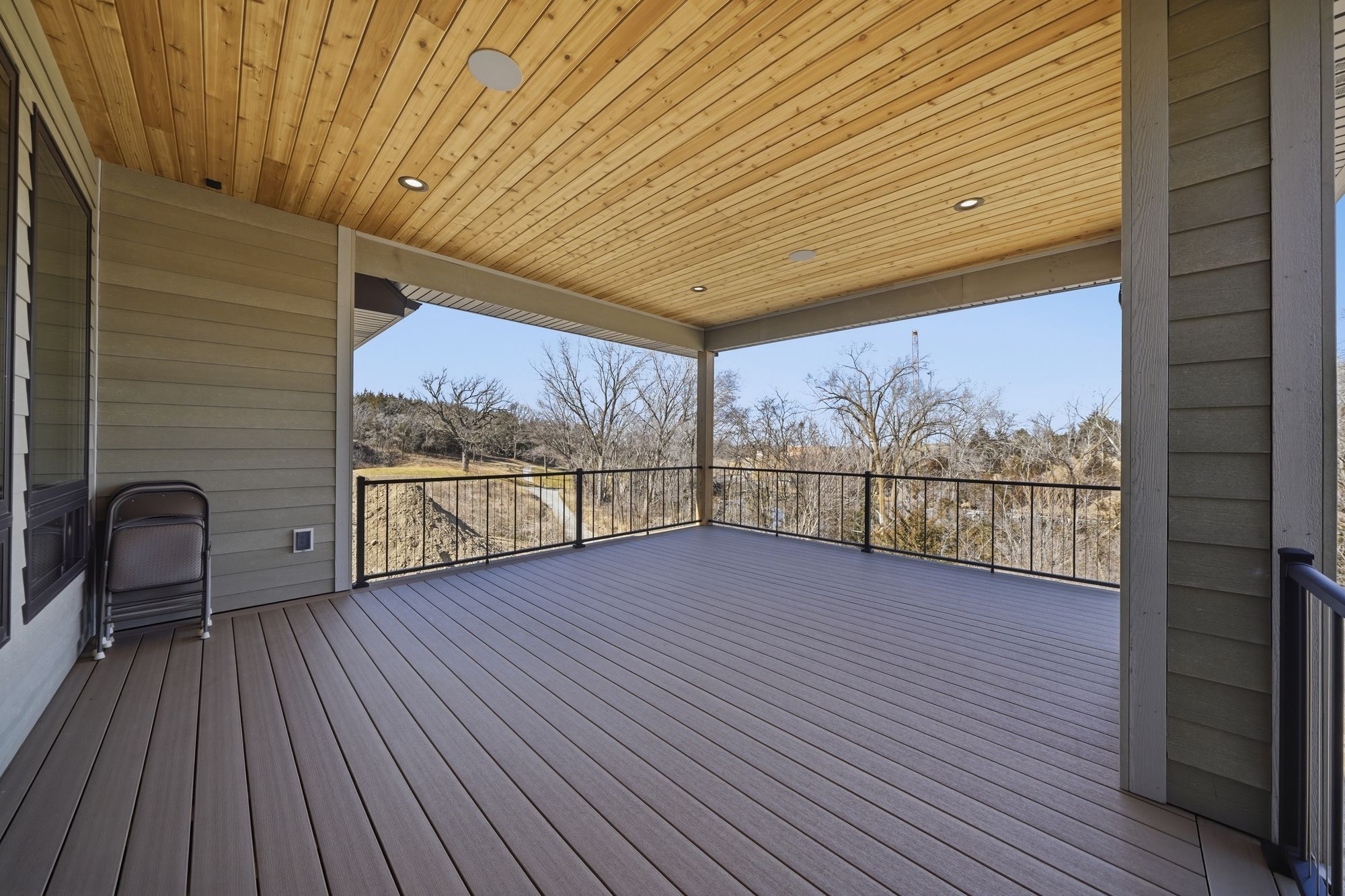 Covered outdoor deck with tongue and groove wooden ceiling and rod iron railing overlooking wooded landscape. Decking is composite plank material.