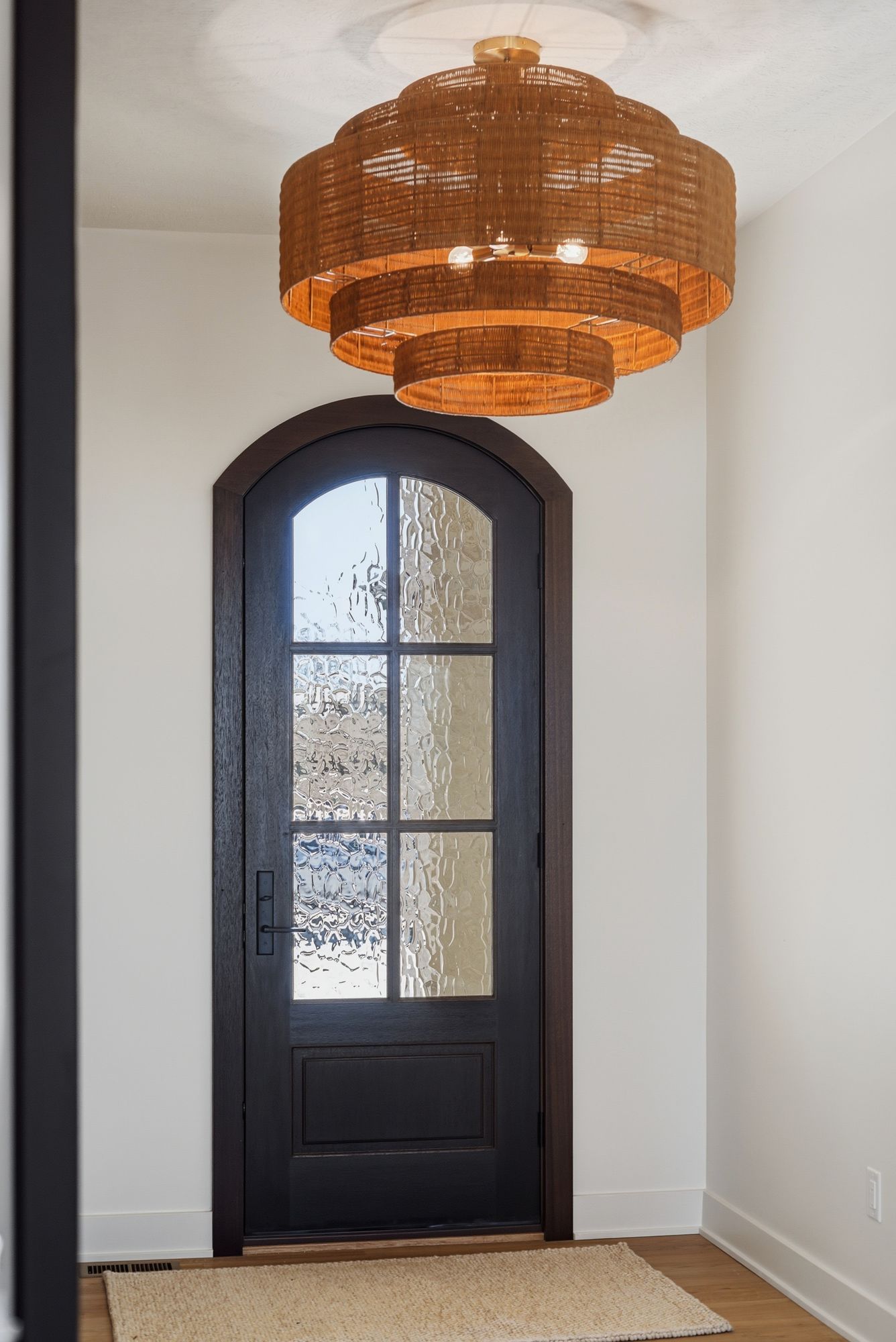 Transitional entry features a dark wood arched door with textured glass, complemented by a tan jute rug and a multi-tiered woven chandelier. The walls are painted in a soft white.