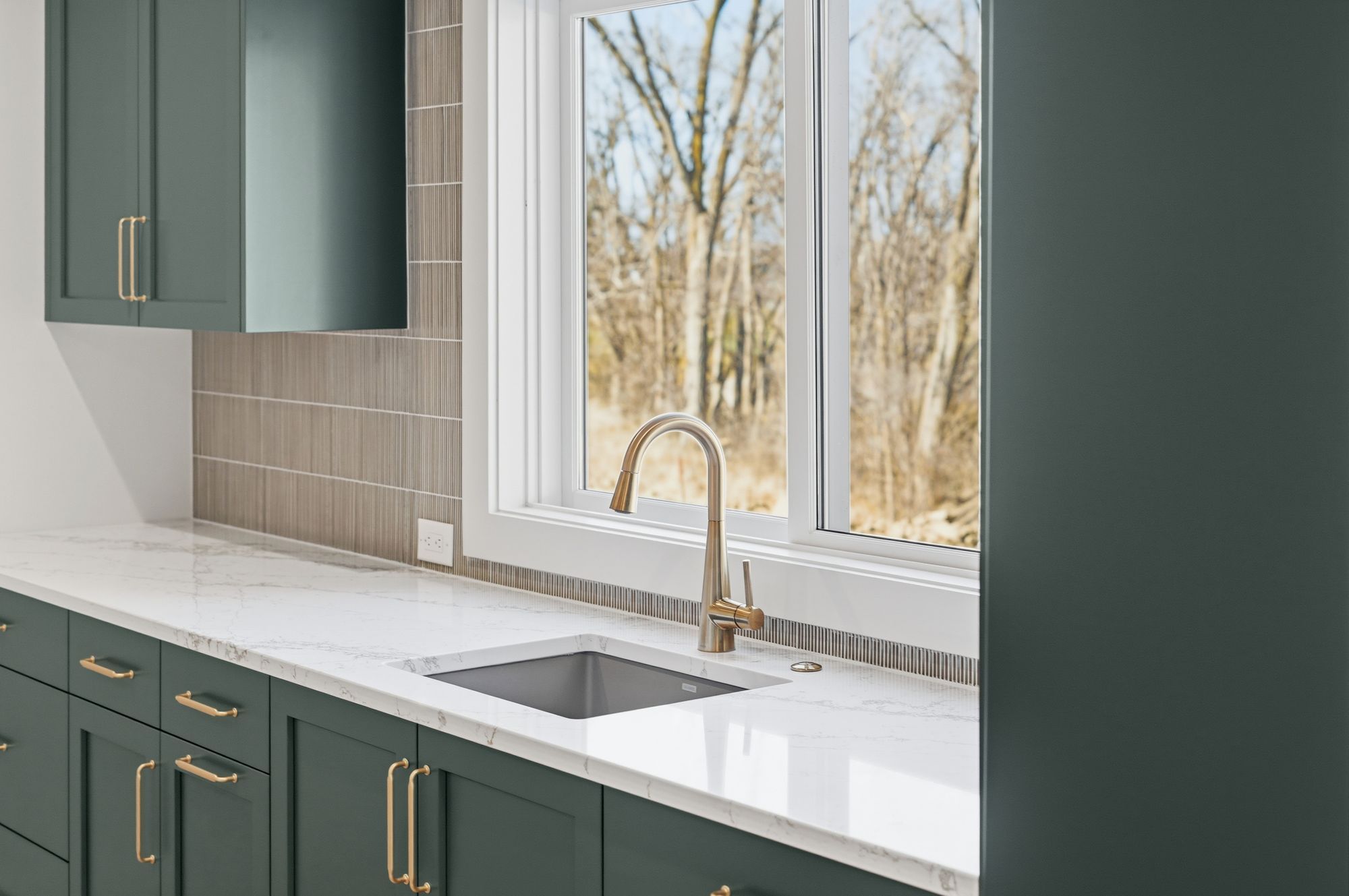 Basement wet bar features dark green cabinets, white countertops, and a stainless steel sink under a window overlooking trees. The design includes gold hardware, a tile backsplash, and modern fixtures.