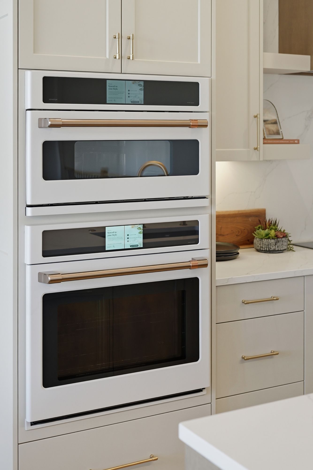 Kitchen showcasing built-in white Cafe appliances, including a convection oven and microwave, complemented by gold hardware and light gray cabinetry. A marble backsplash and countertop add a luxurious touch to the space.