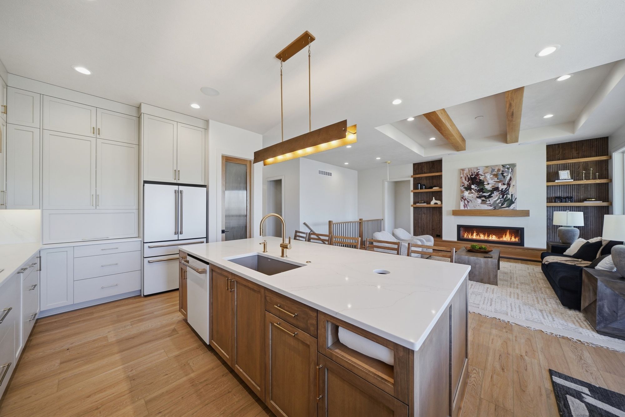 Modern kitchen with light wood floors features a large island with a white countertop and wood cabinets. It opens into a living room with a fireplace and built-in shelving.