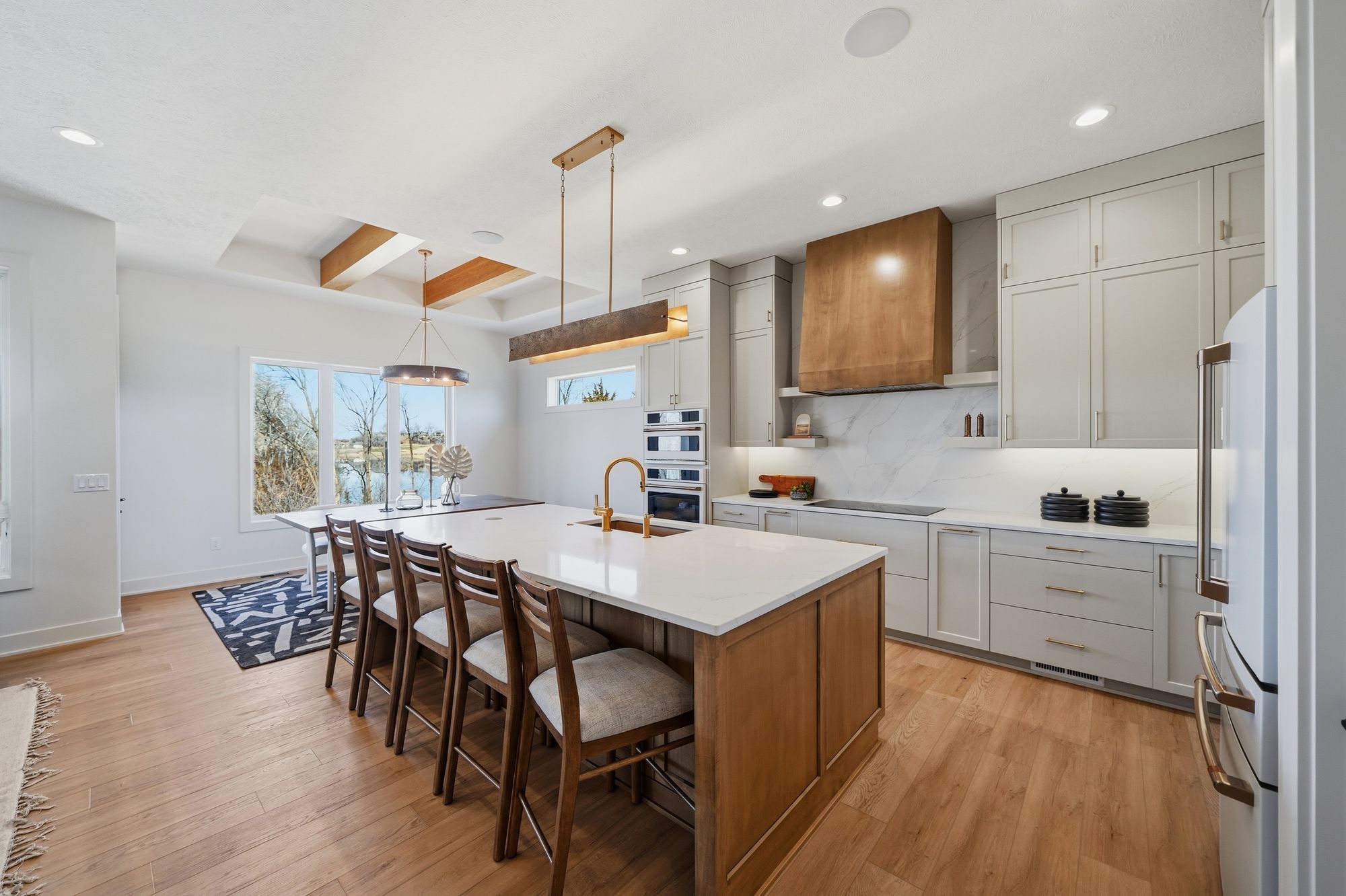 Bright kitchen features a large island with seating, quartz countertops, a gold faucet, and light gray cabinetry. There is a copper range hood and wood flooring throughout the space.