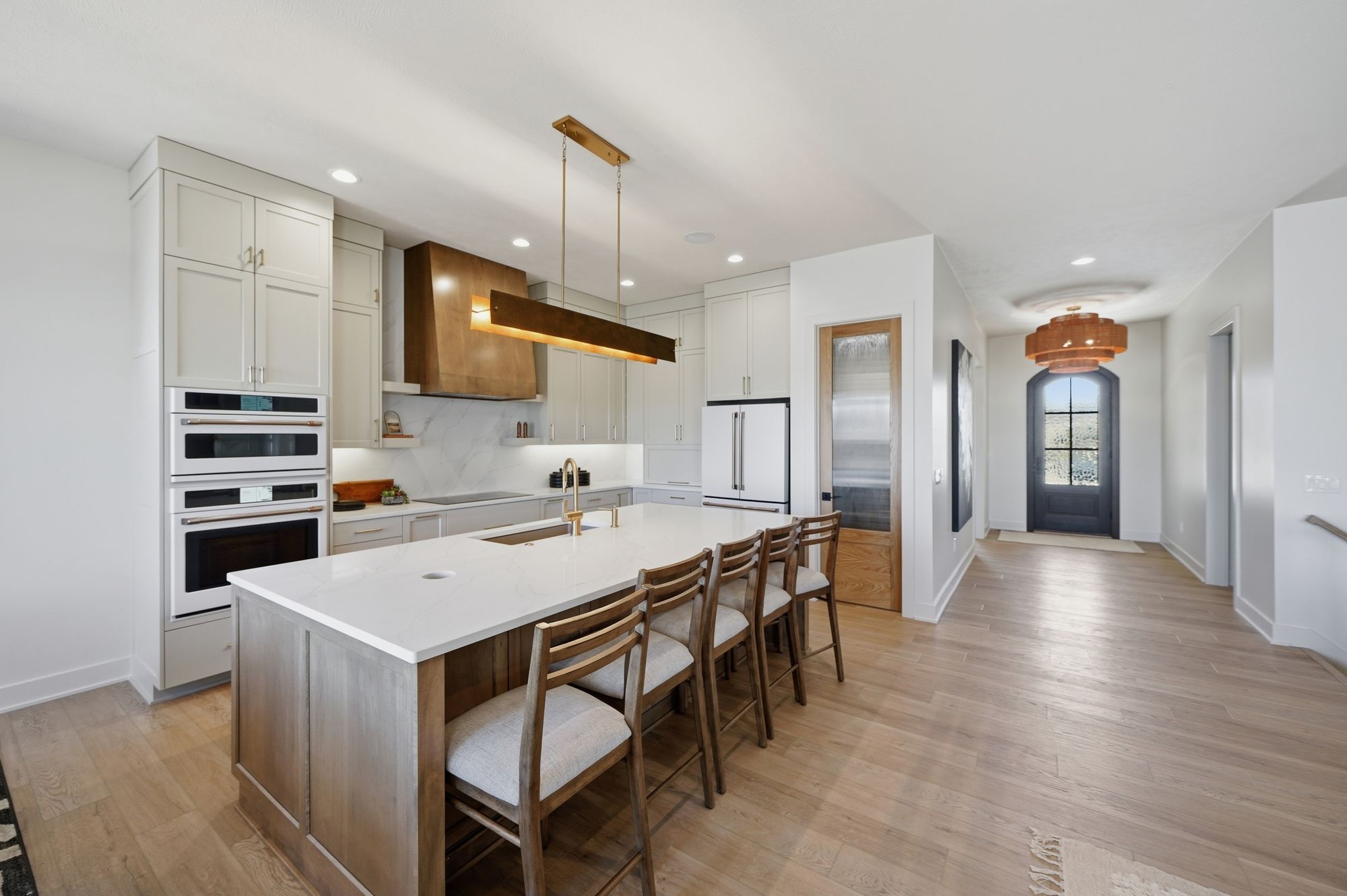 Bright, modern kitchen features white cabinets, marble countertops, and a large island with seating. The space opens into a hallway with a front door visible, creating an open and airy atmosphere.
