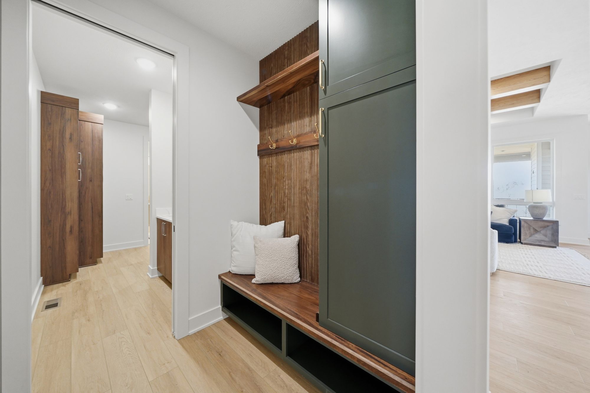 Interior mudroom featuring a built-in bench with open storage below, a wood-paneled back, and a tall green cabinet. The room opens to a hallway with wood cabinets and a view to a living area with a blue chair and white rug.
