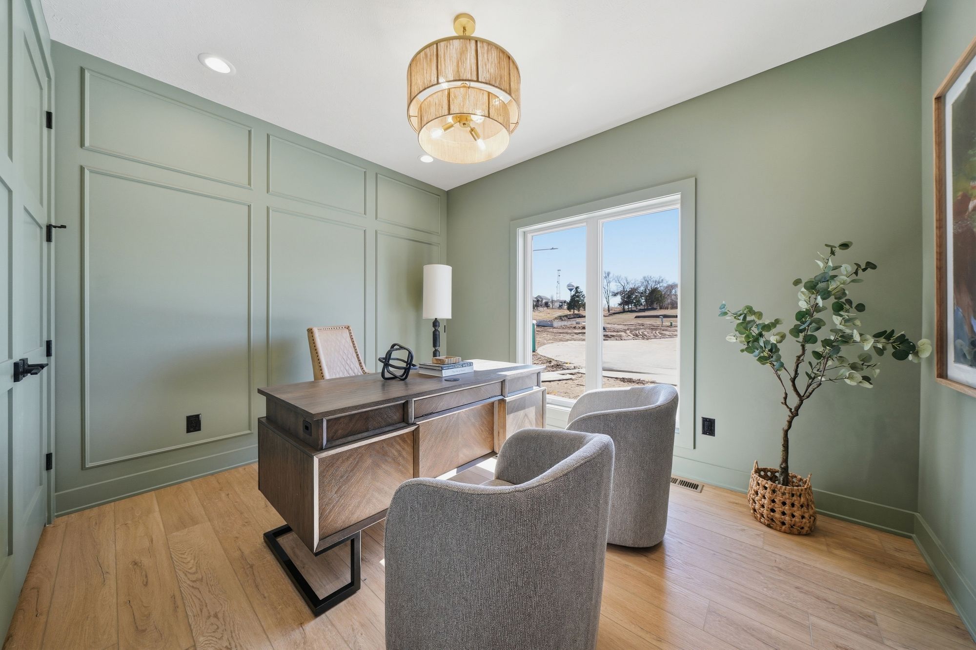 Office space with sage green walls, wainscoting, and wood floors features a contemporary desk with an interesting base. Two gray upholstered chairs face the desk and natural light comes through a window overlooking a construction site.