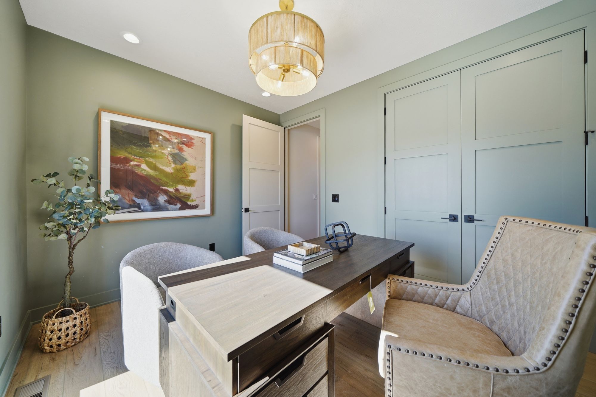 This is an office space with two chairs, a wooden desk, and olive green walls. It features a large framed painting, a decorative tree in a basket, and a modern light fixture.