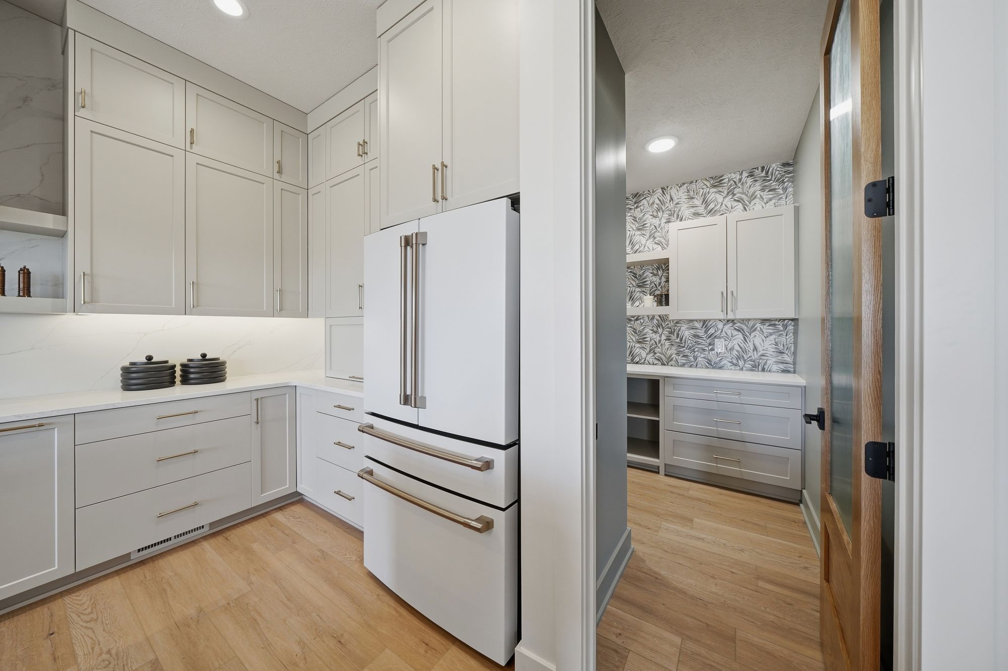 Bright pantry with white cabinetry, countertops, and appliances complemented by wood-look flooring. The adjacent room features leaf-patterned wallpaper and matching built-in storage.