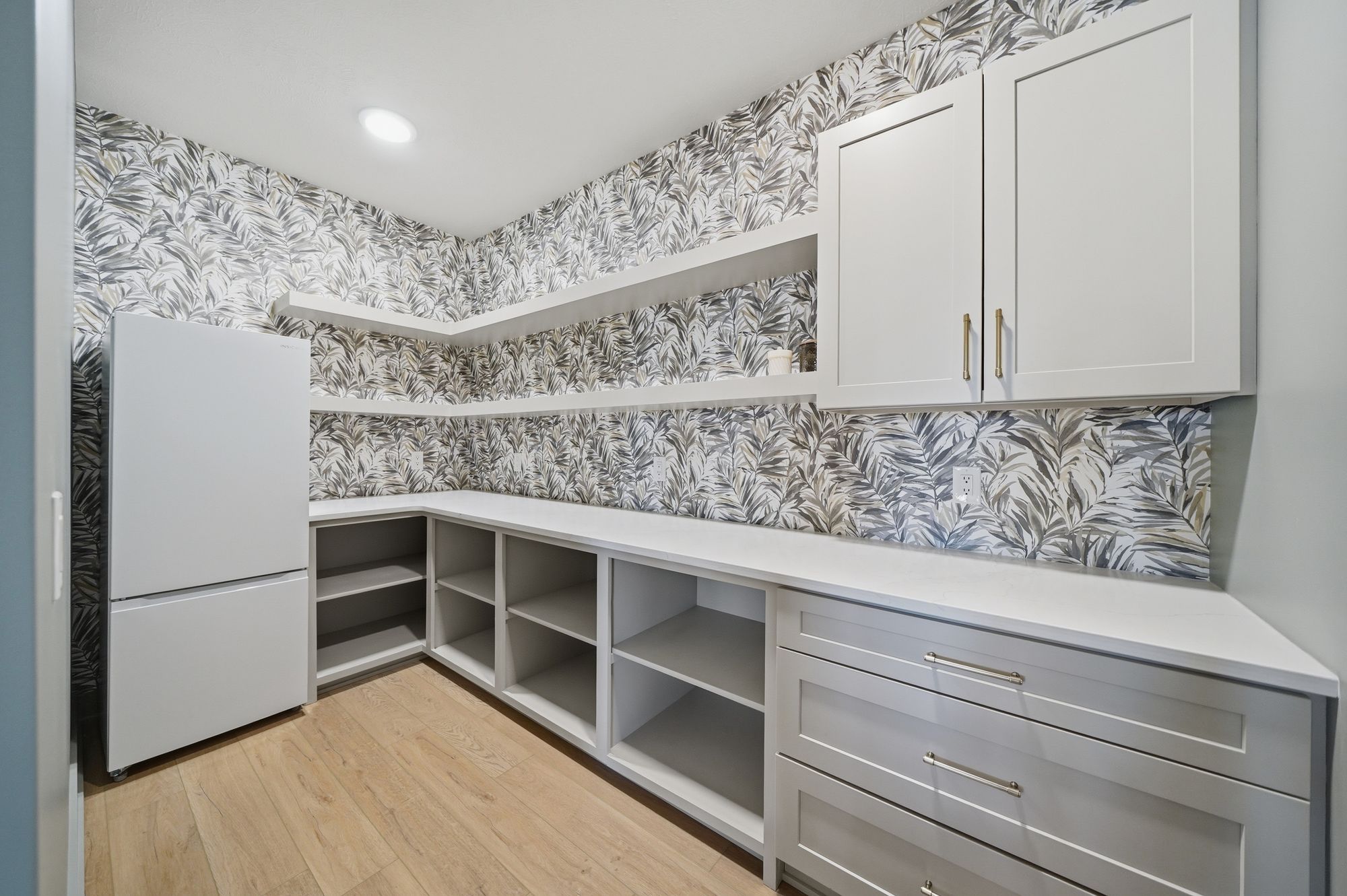 A well-organized pantry features a white refrigerator, white cabinets with brushed gold hardware, open shelving, and drawers, all accented by leaf-patterned wallpaper and light wood-look flooring.