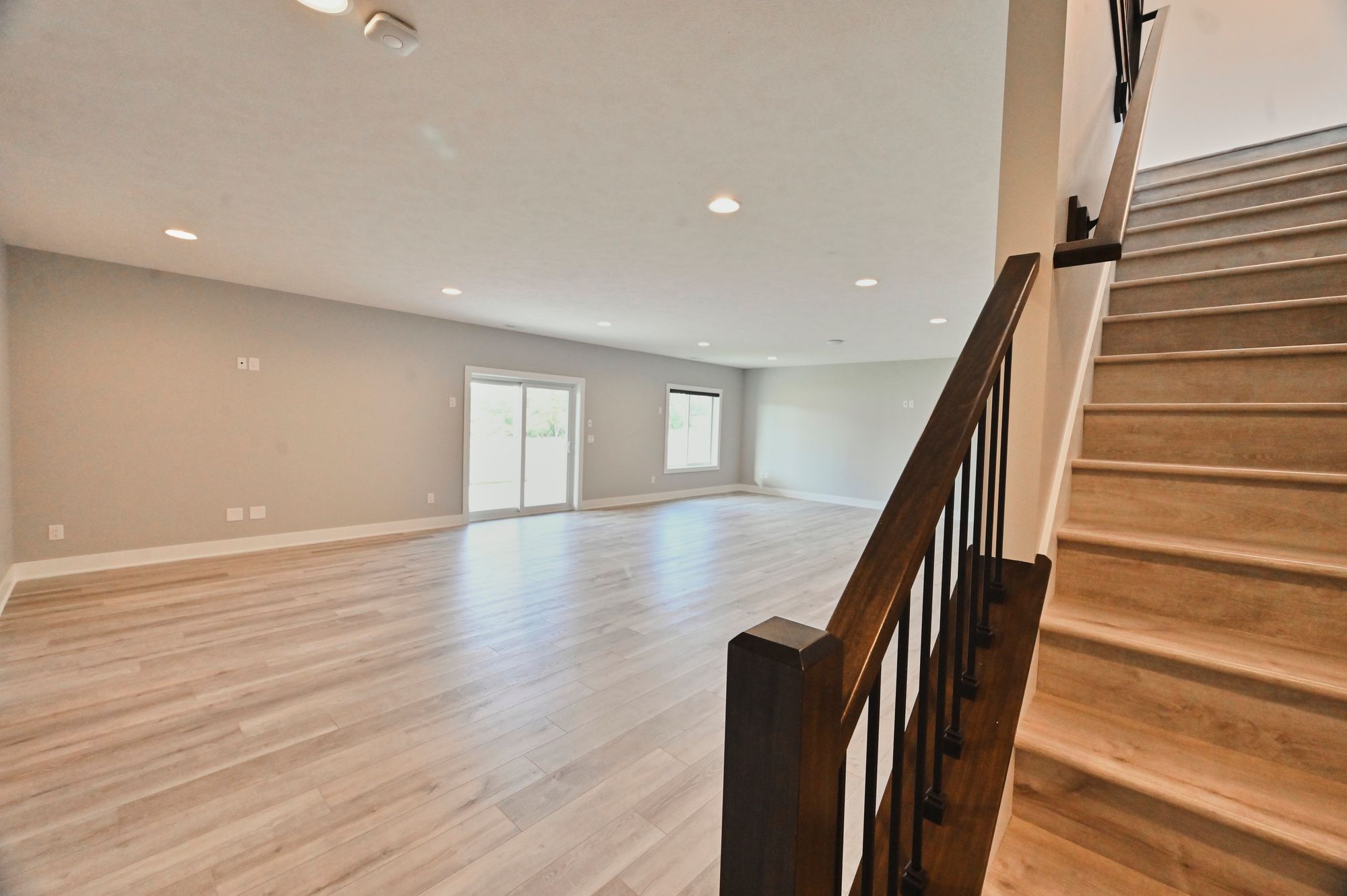 Spacious basement with light wood-look flooring, sliding glass doors to the backyard, and a window for natural light. Wood stairs with dark metal railing lead to another level.