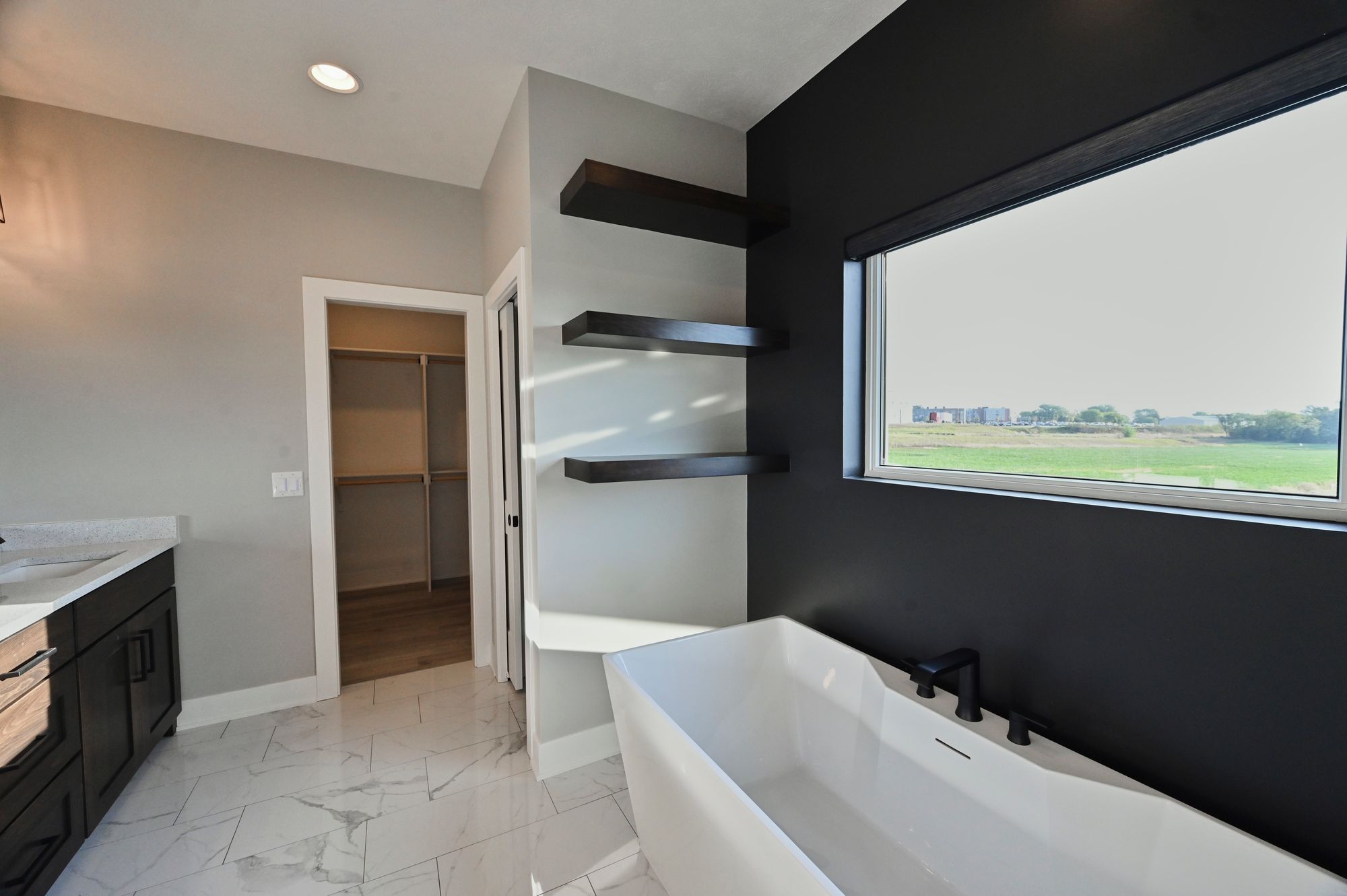 Modern bathroom with a freestanding tub, dark accent wall, and floating shelves is shown. A large window provides natural light and a view of the outdoors, and marble-look flooring is featured.