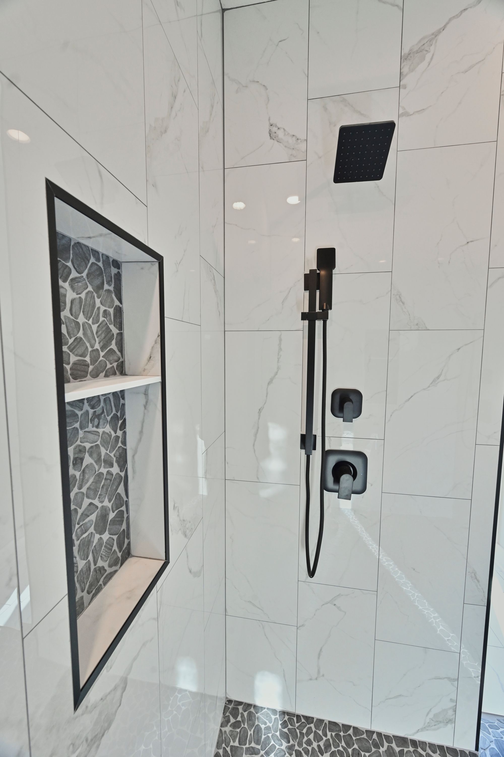 Modern bathroom shower with black fixtures, including a rain shower head, a handheld shower wand, and square control valves. A built-in niche with a black frame and pebble tile accents provides shelving.