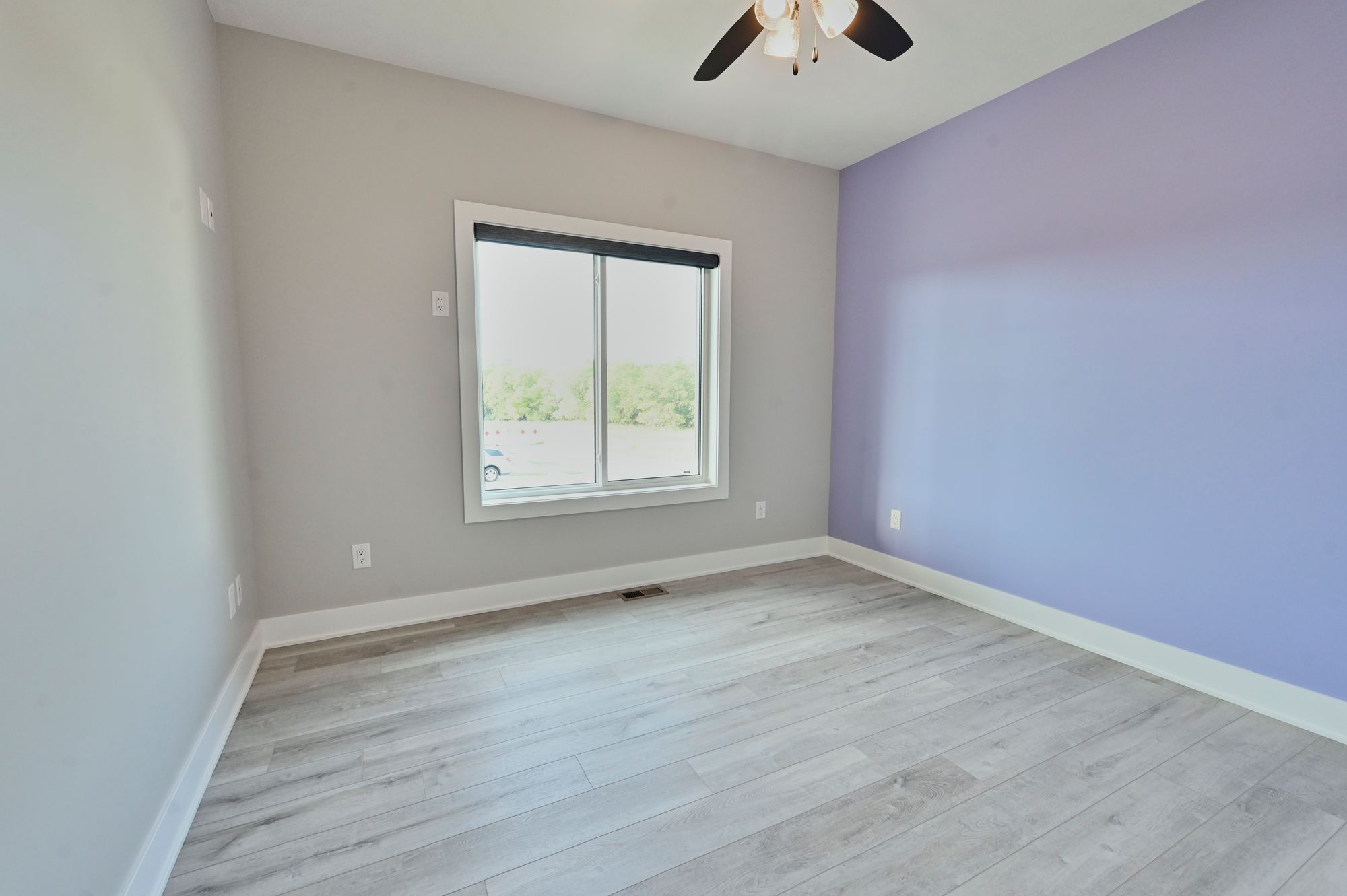 This is a bedroom with light gray laminate flooring, light gray walls, and one wall is light purple. There is a window with a black shade.