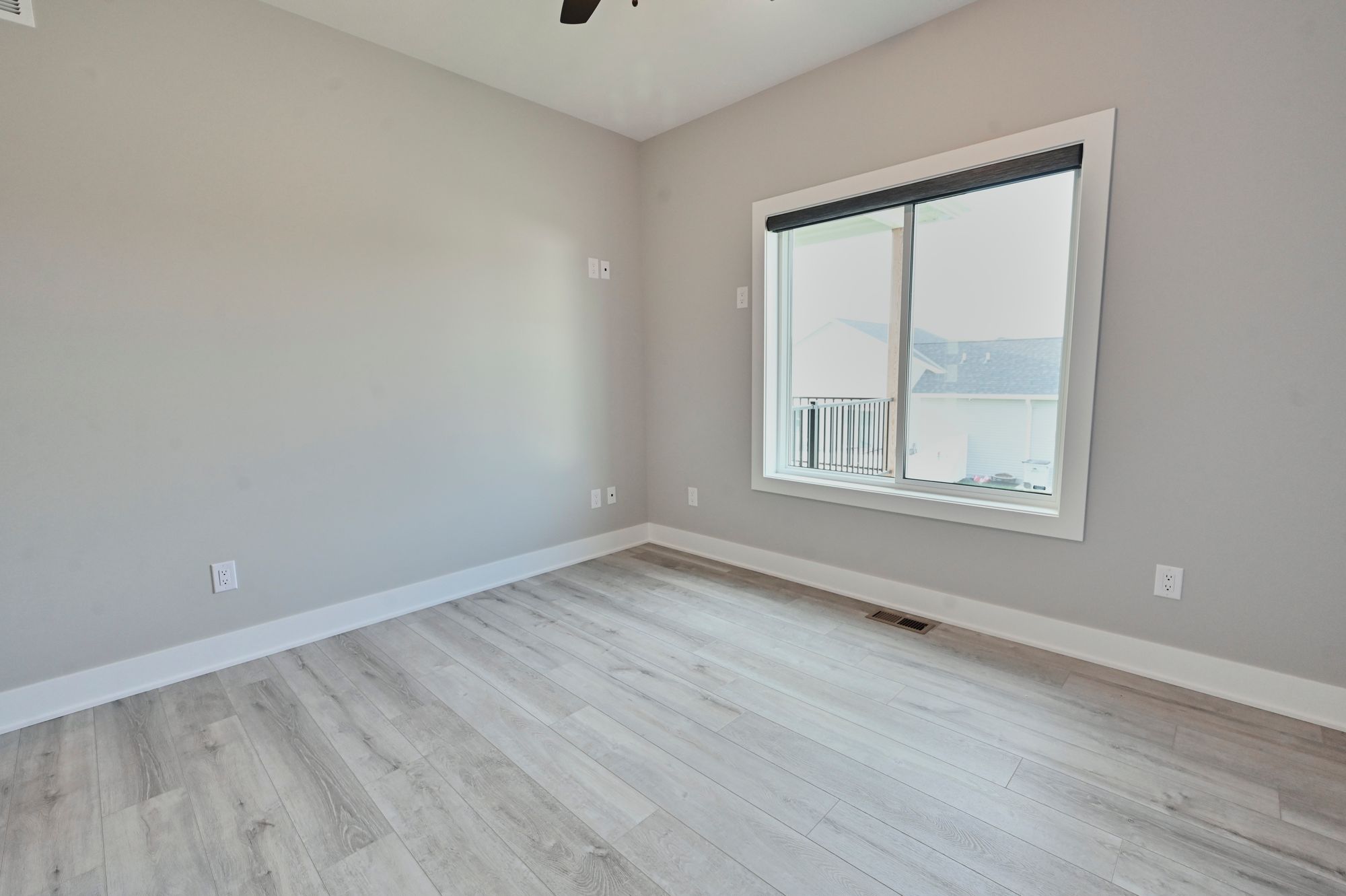 A bedroom features light gray walls and light wood laminate flooring. There is a large window with a white frame and sliding glass door, offering a view to the outside.