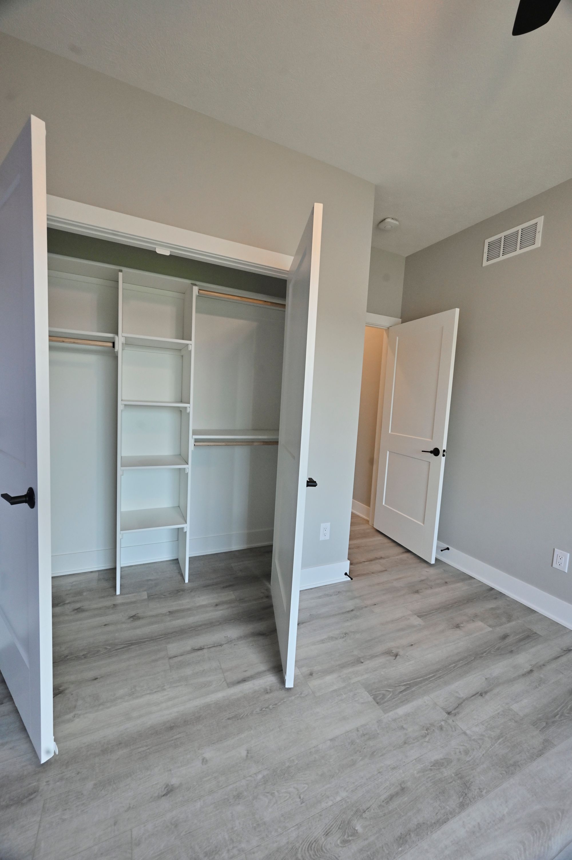 The image showcases a bedroom featuring a closet with white shelving and doors, and light gray walls paired with light gray wood-look flooring. The closet door is ajar, revealing the interior, while a second door leads out of the room.