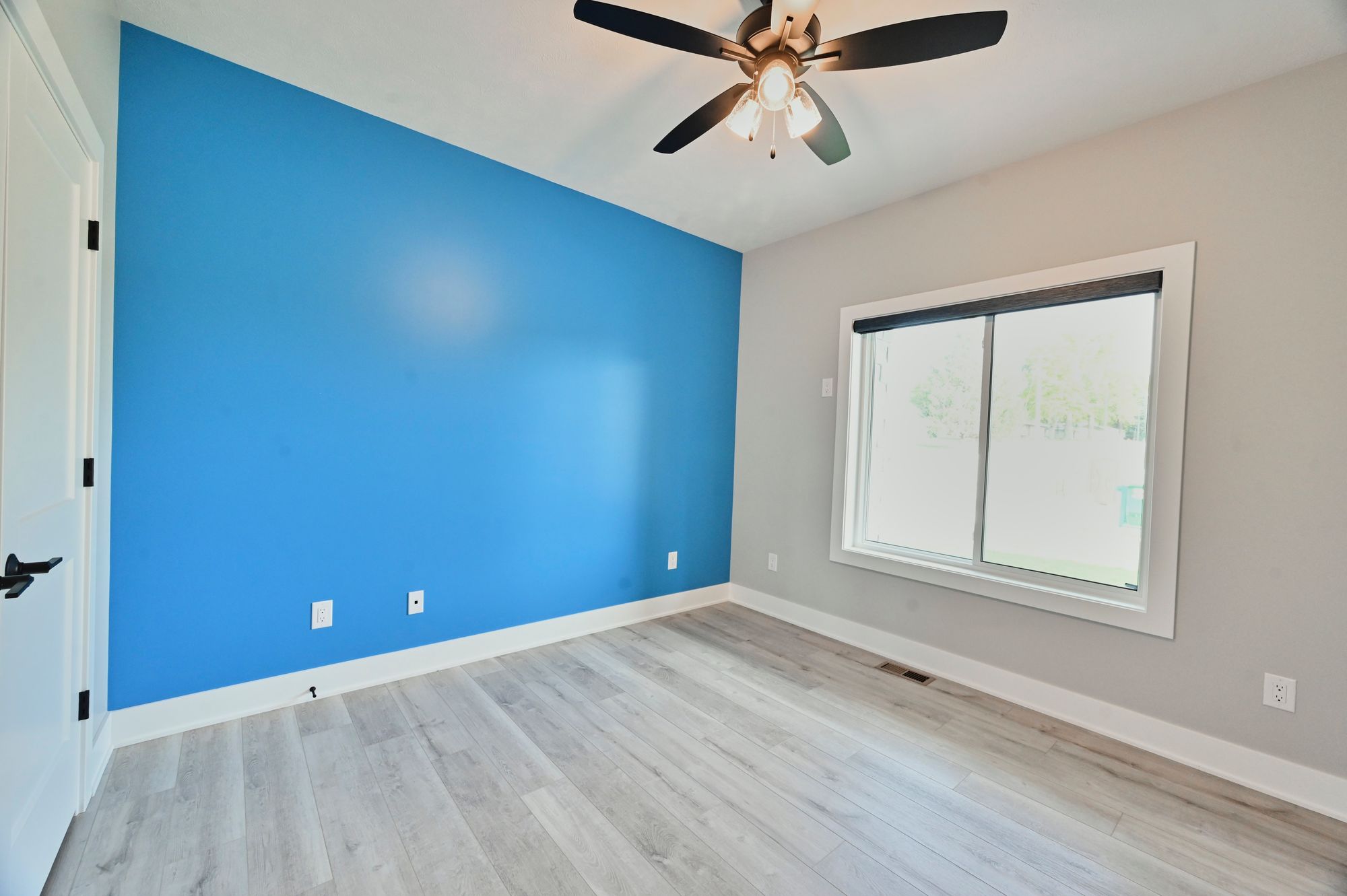 Bright bedroom features a bold blue accent wall, light gray walls, and light gray hardwood floors. The room also has a ceiling fan and a double-hung window with a white frame.