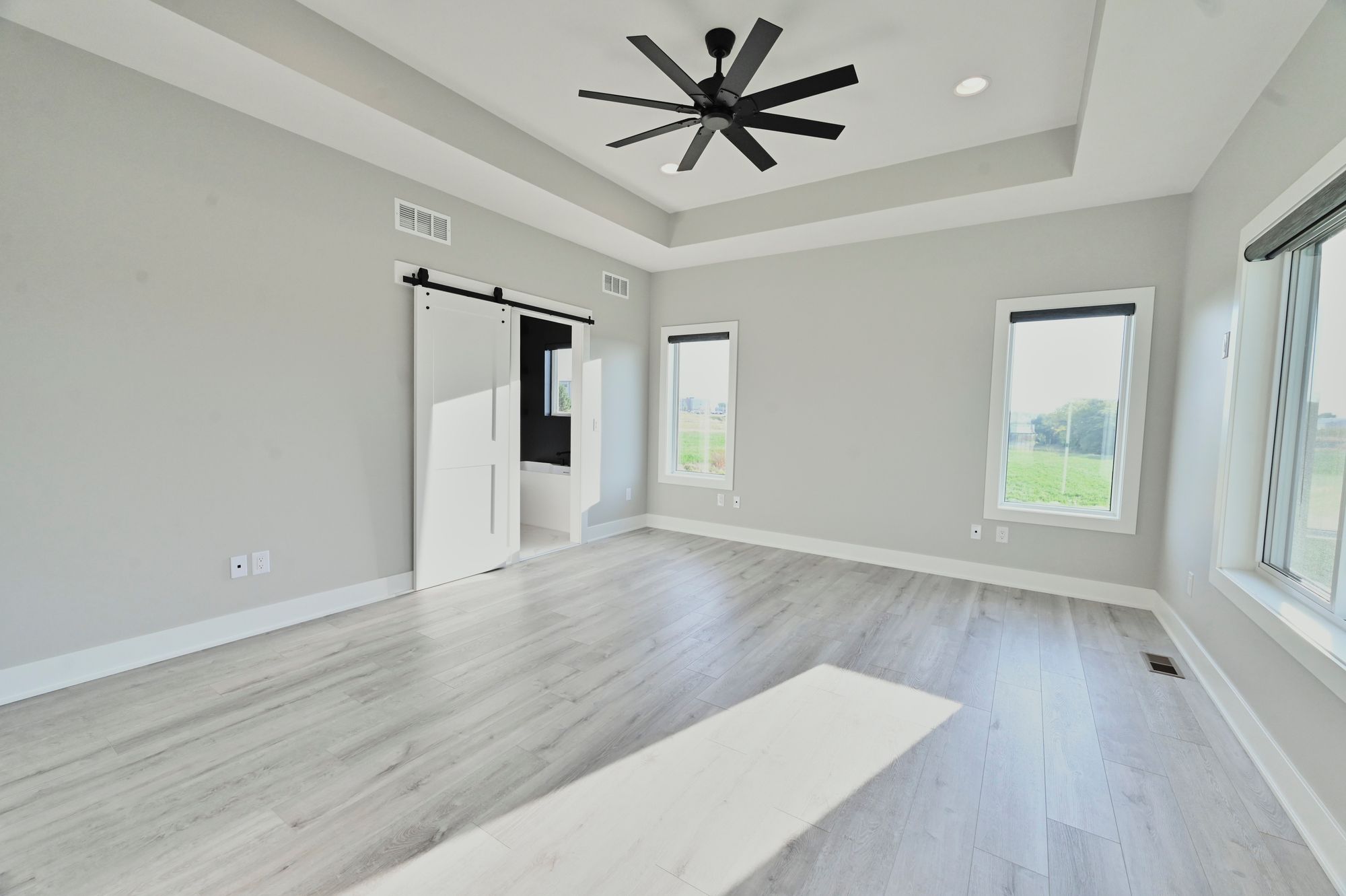 The bedroom features gray walls, light hardwood flooring, and a modern barn door leading to the bathroom. There's a ceiling fan and a double window allowing natural light.