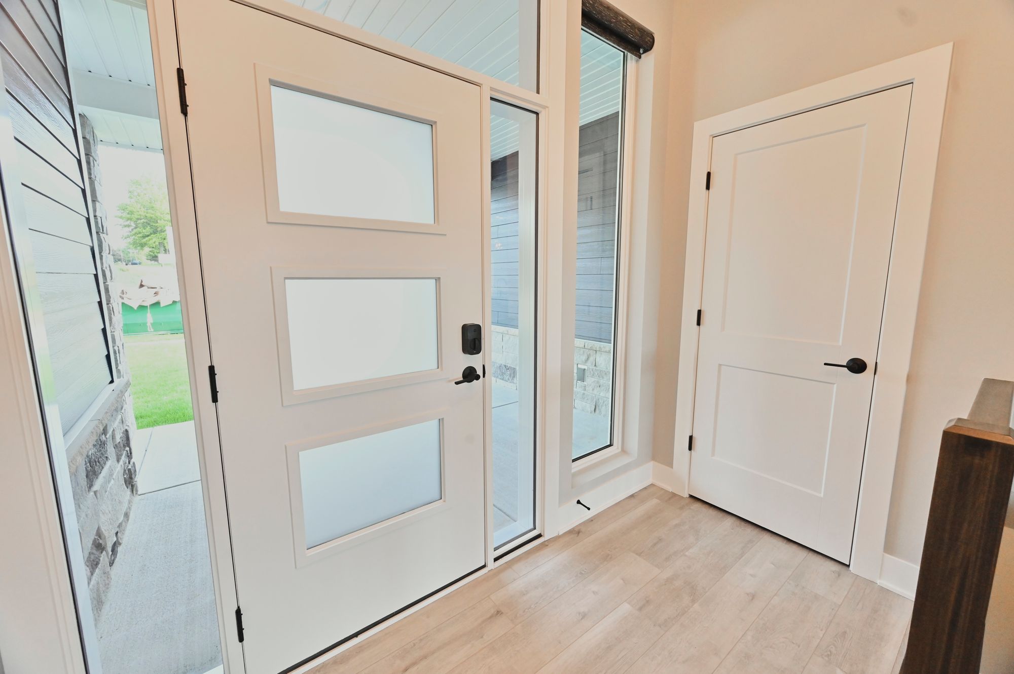 A modern entry features a white front door with three frosted glass panels, complemented by a narrow sidelight window. Inside, light wood floors lead to a white interior door with black hardware.