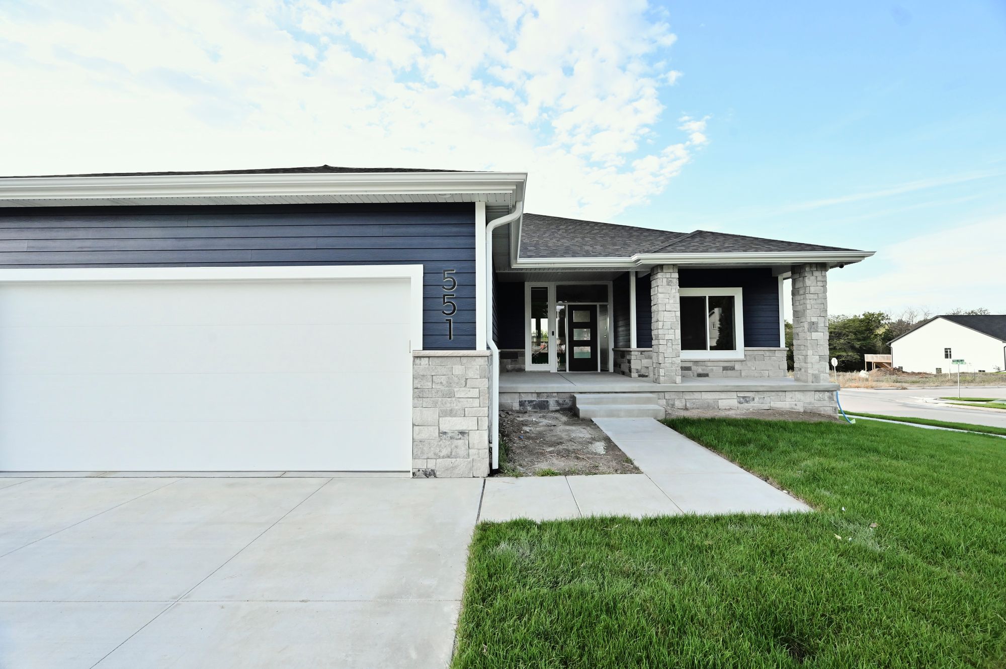 Exterior view of a one-story home featuring dark blue siding, stone veneer accents, and a white garage door. A concrete driveway leads to the garage, and a walkway extends to the front porch with dark front door.
