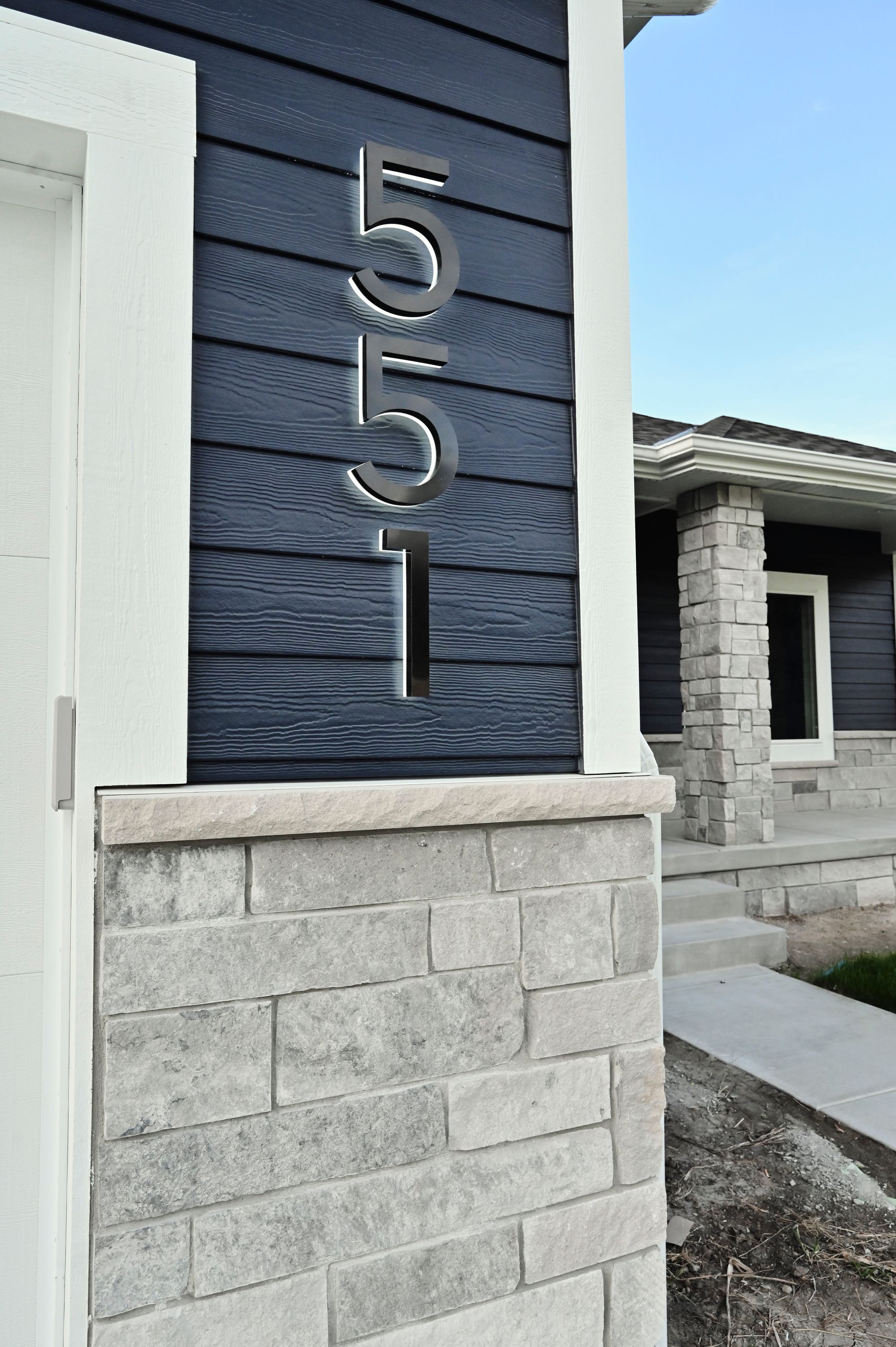 Modern home exterior shows dark blue horizontal siding with large silver house numbers '551'. Stone veneer accents the base, with a white door frame and stone porch column visible.