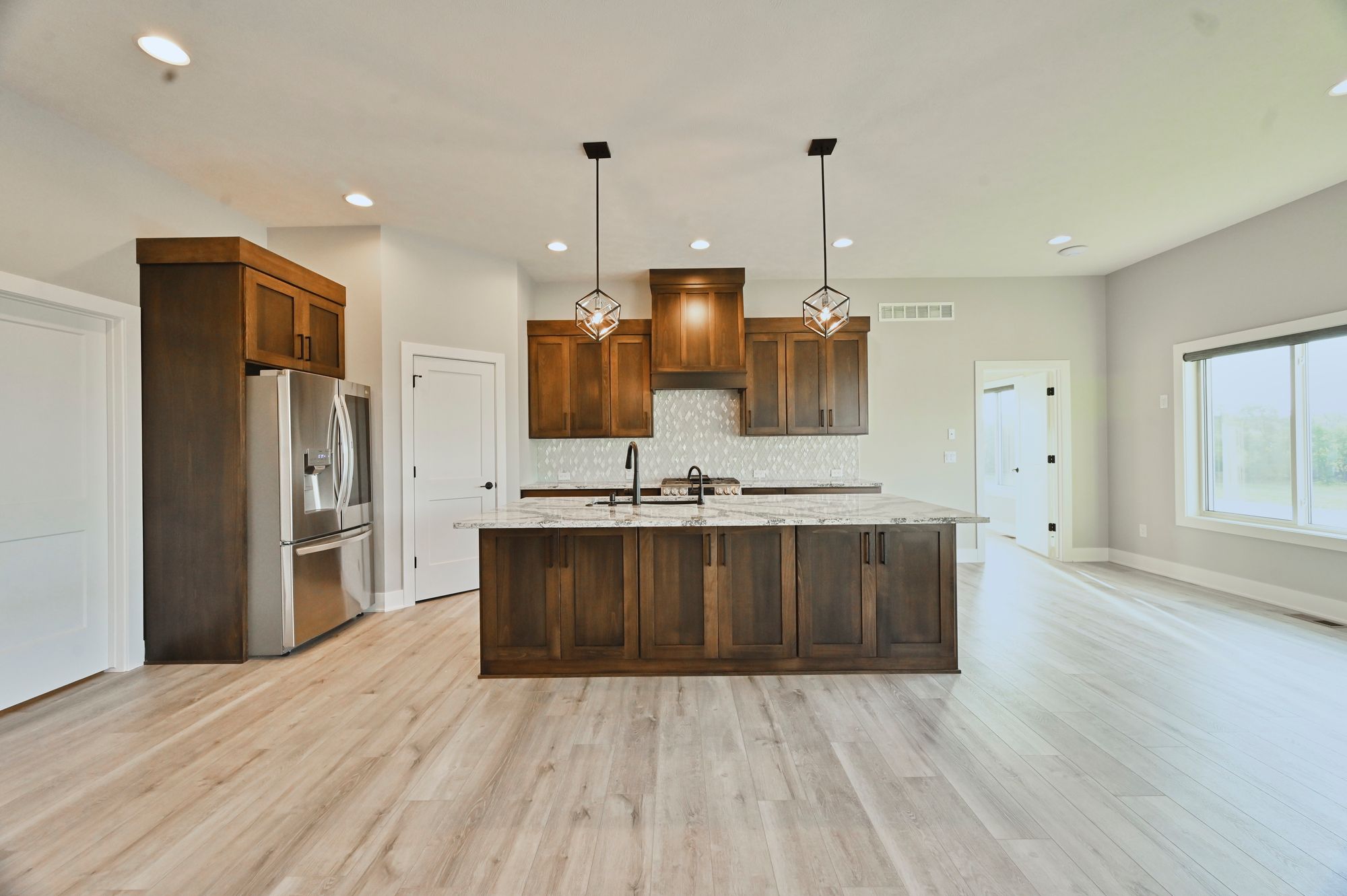 Modern kitchen features stained wood cabinetry, a spacious island with granite countertops, and stainless steel appliances. The flooring is light-toned wood-look plank, and pendant lights hang above the island.