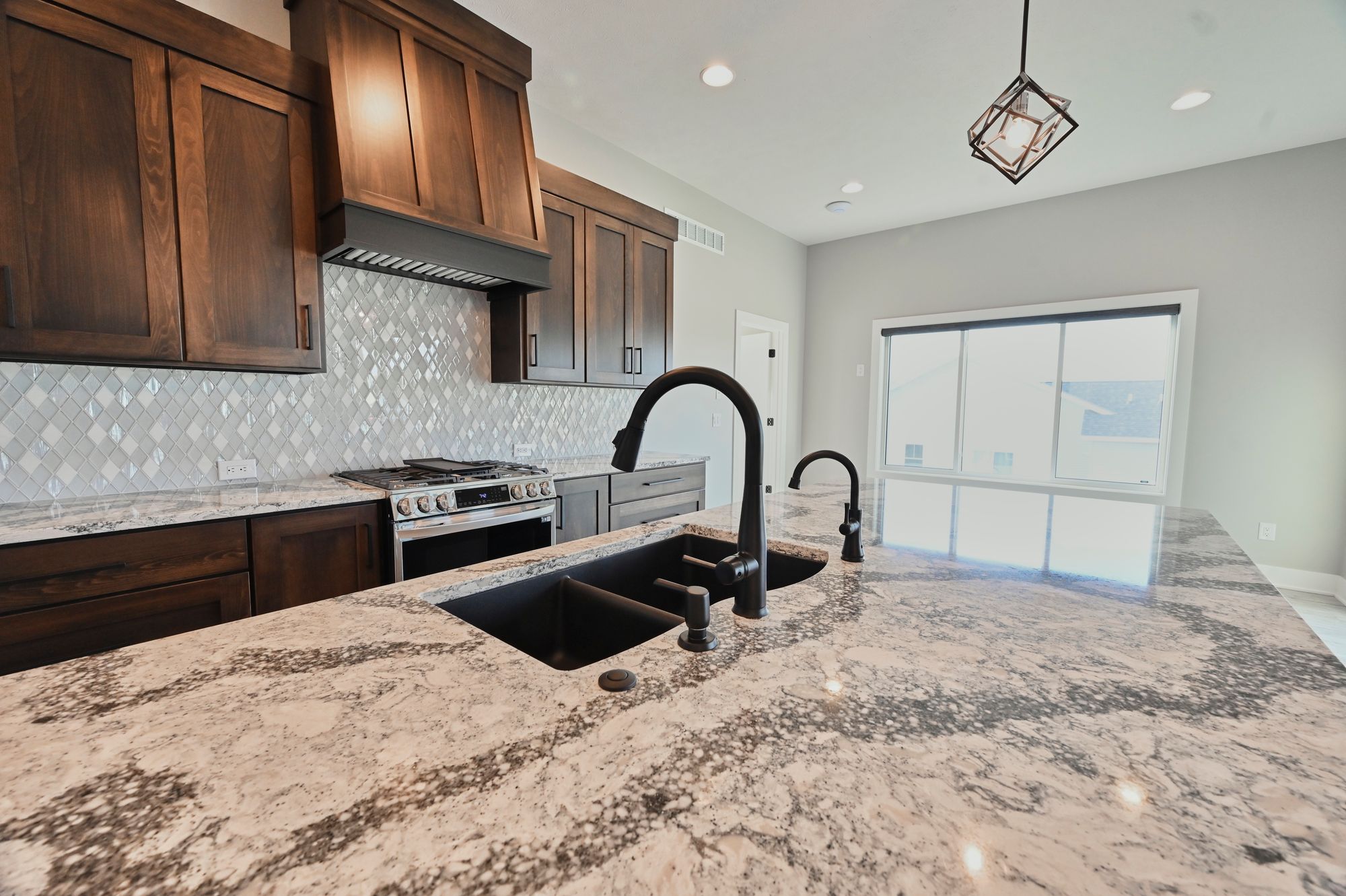 Modern kitchen featuring a large island with a black sink and fixtures, surrounded by speckled granite countertops. Dark wood cabinets are paired with a patterned backsplash and stainless steel stove.
