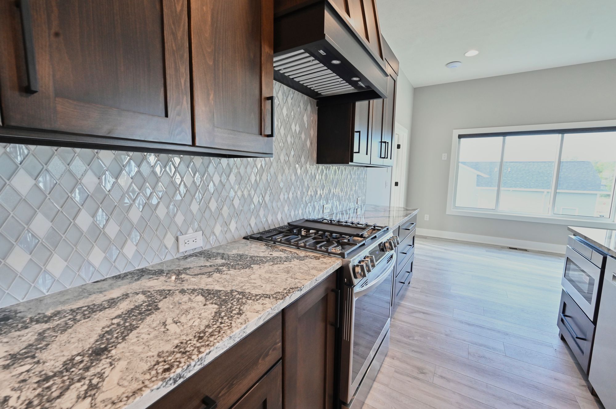 Modern kitchen featuring dark wood cabinetry, granite countertops, and stainless steel appliances including a gas range. The backsplash is light gray diamond patterned tile.