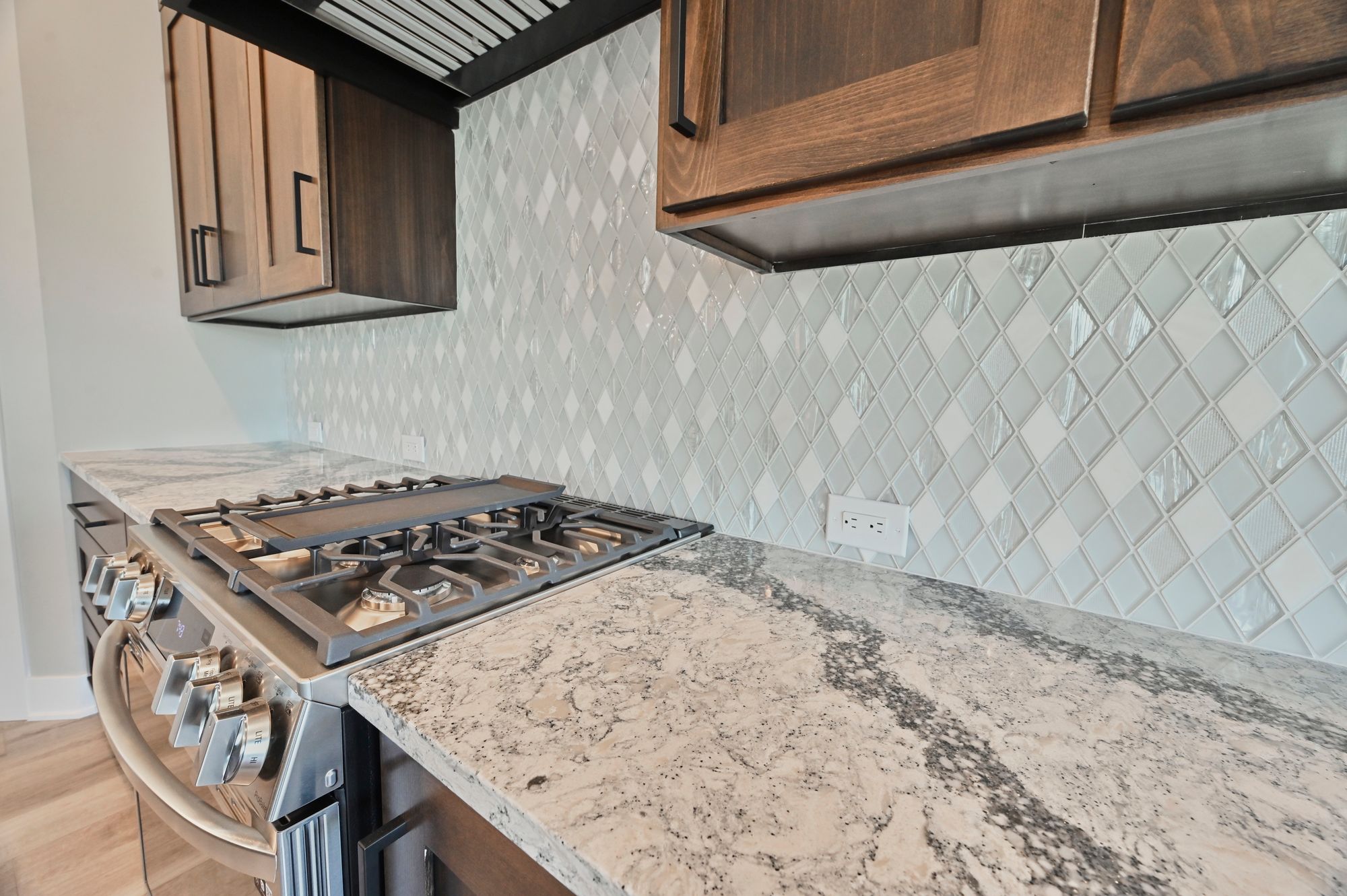 Kitchen showcases stainless steel gas range, granite countertops, and light gray diamond-patterned backsplash. Dark brown cabinets are above the range, adding a touch of warmth.
