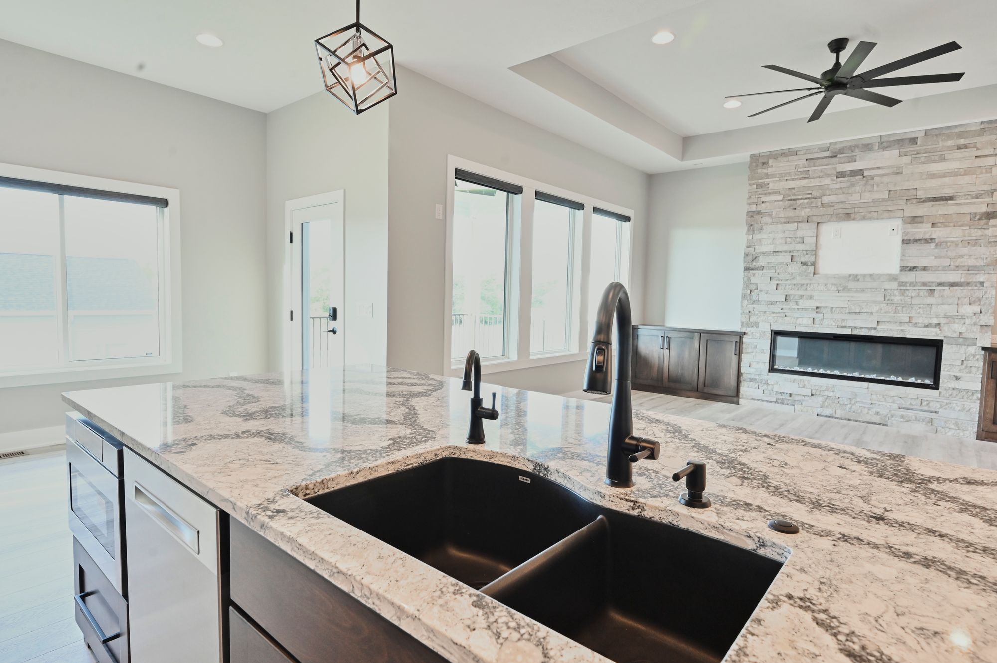 Modern kitchen features a granite countertop island with a black undermount sink and sleek black faucet. The kitchen opens to a living area with a stone fireplace and large windows.