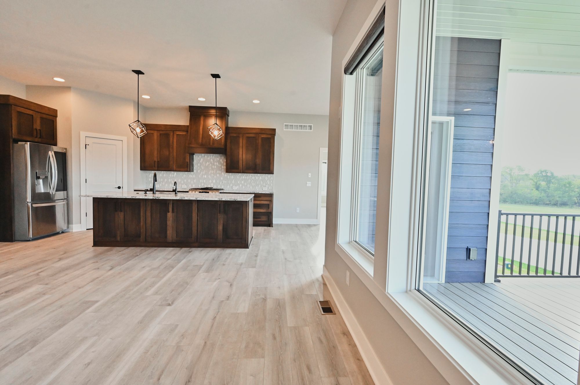Open concept kitchen features dark wood cabinetry, a large island with white countertops, and stainless steel appliances. The space opens to a covered deck with gray railings.