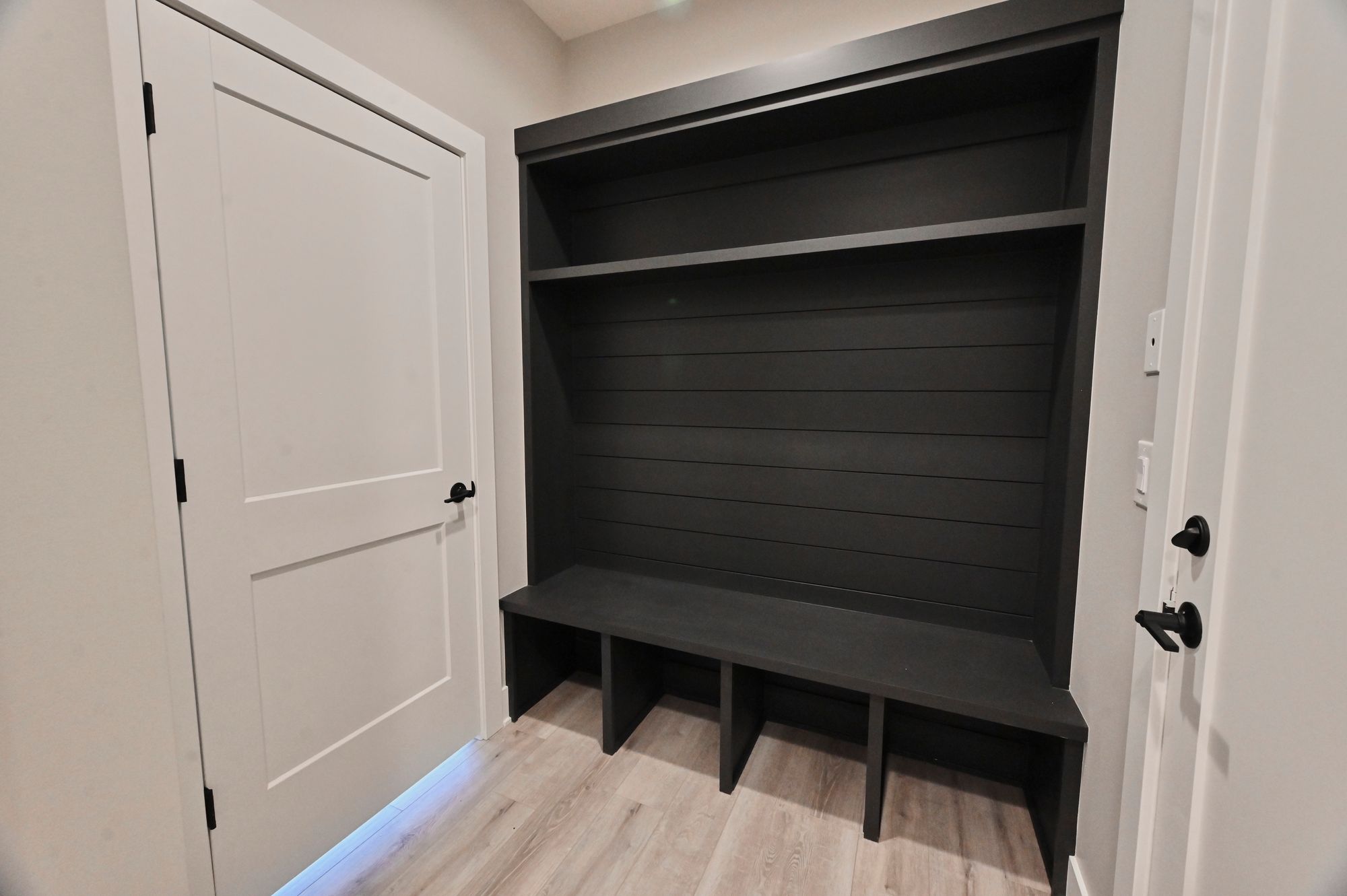 Mudroom with built-in dark gray storage bench and shelving, located next to a white door. The flooring is light wood.