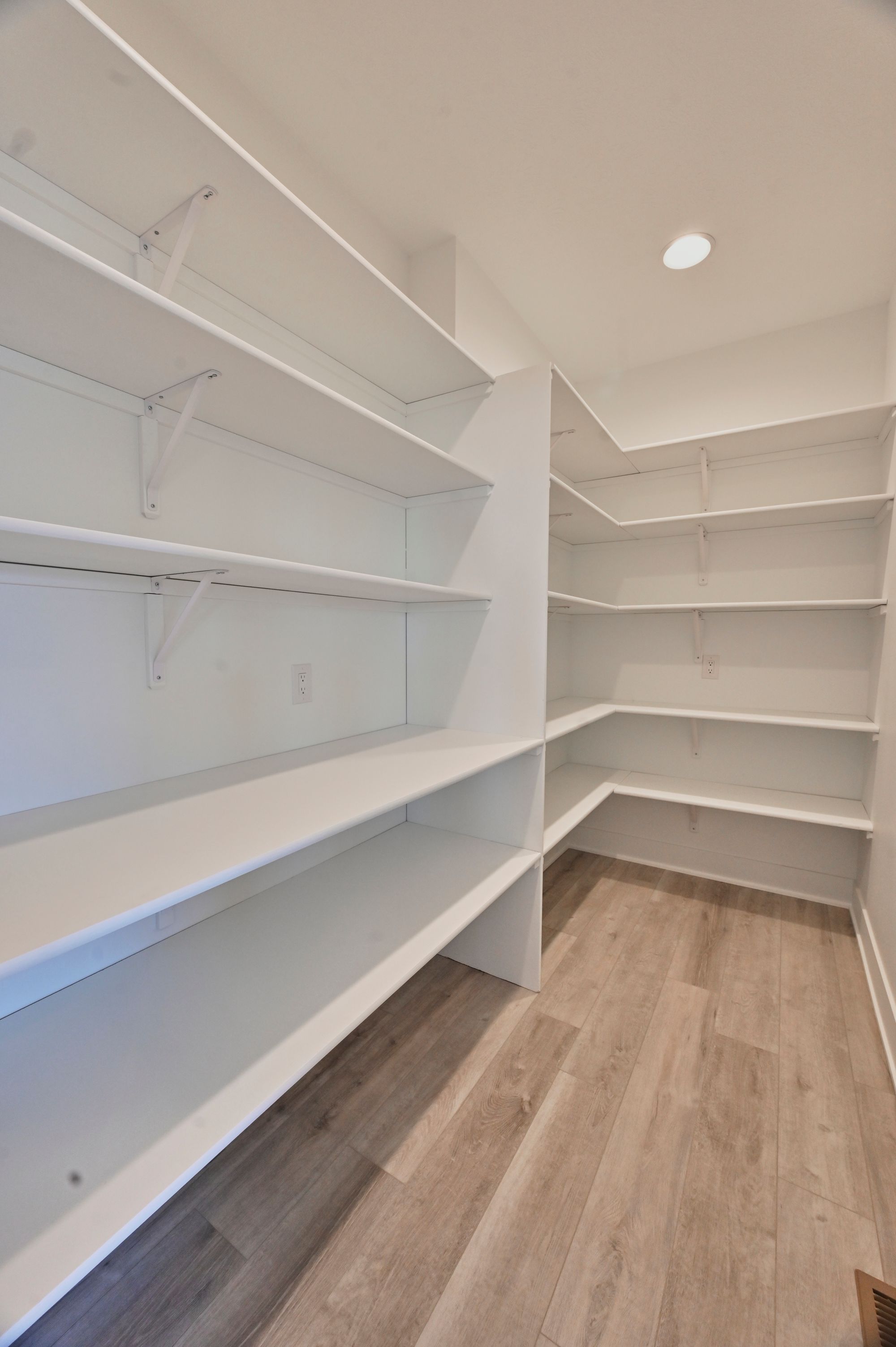 The image showcases a well-lit pantry with built-in white shelving on multiple walls and a light wood-look floor. The shelves are empty, and an electrical outlet is visible on one wall.