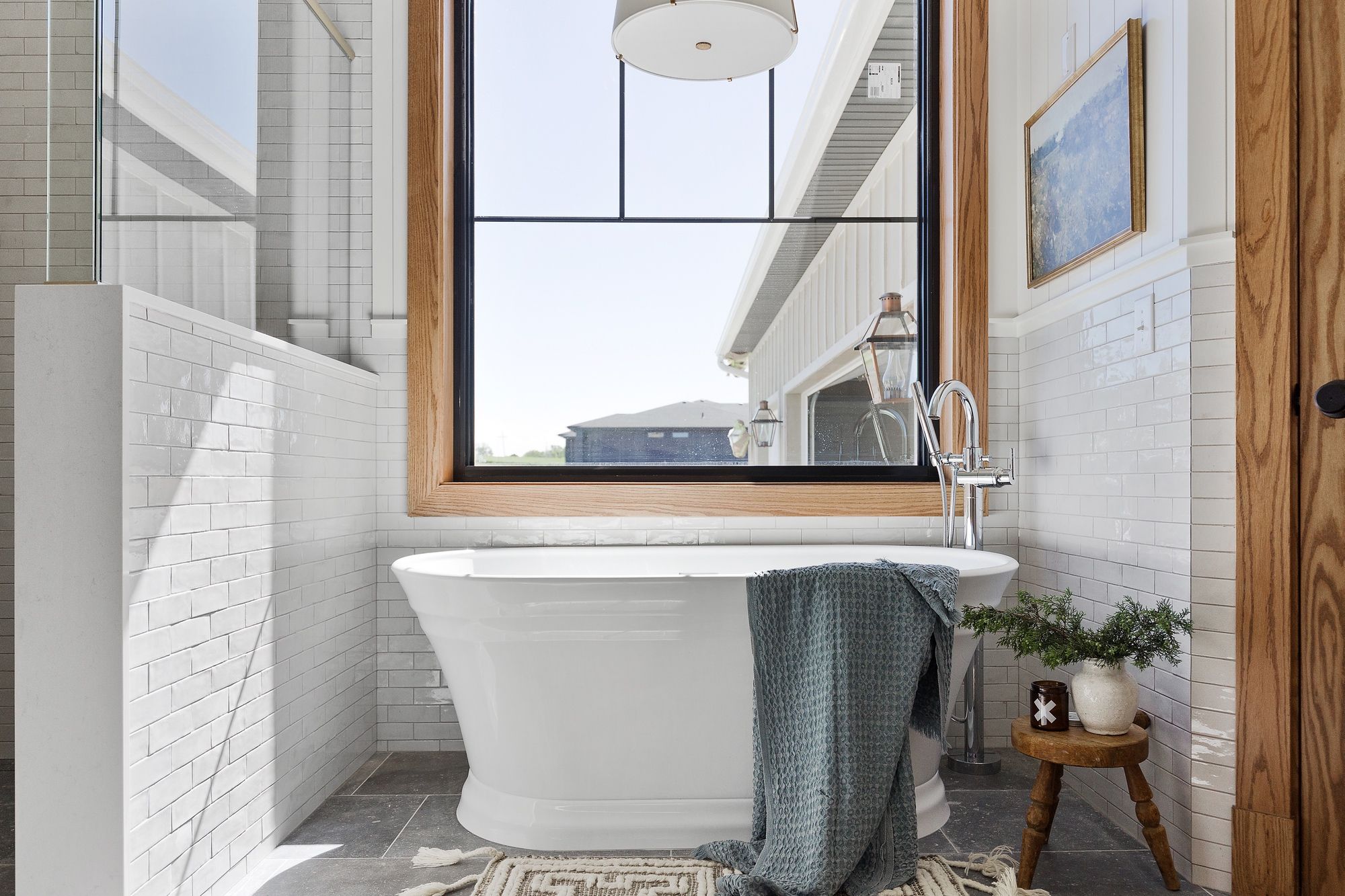 Bright bathroom features a white freestanding tub positioned beneath a large window with black mullions and wood frame. The bathroom also has white subway tile, a dark gray tile floor, and a small wooden stool with greenery next to the tub.
