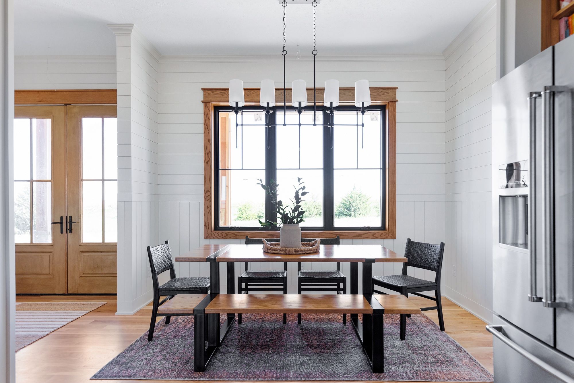 Dining room featuring a wooden table with black metal legs, surrounded by black woven chairs and a wooden bench. The room has white shiplap walls, a large window with a wooden frame, and a modern chandelier.
