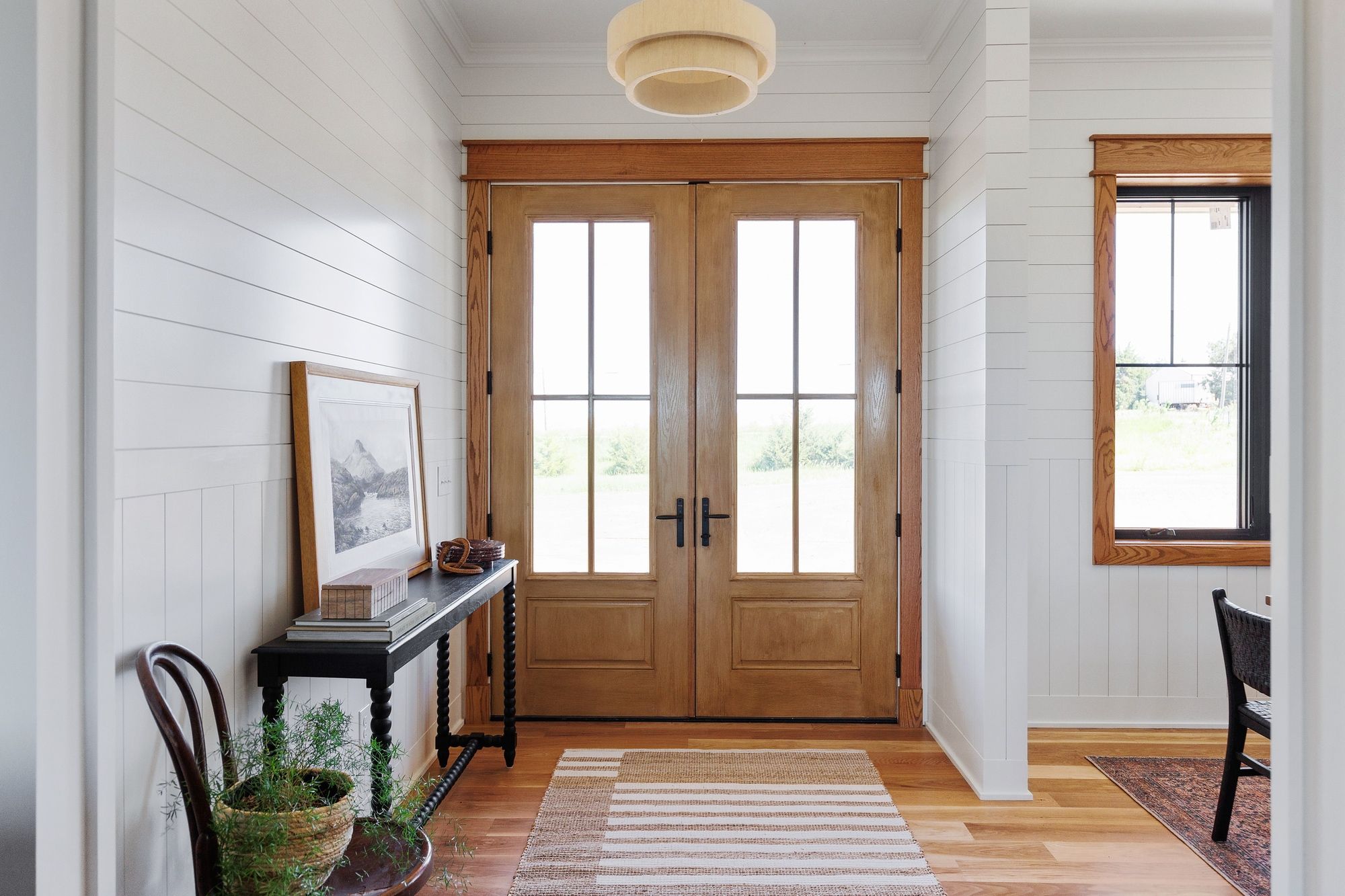 Entryway featuring double doors, shiplap walls, and hardwood floors. A console table with a framed picture and woven accents sits to the left, while a striped rug leads towards the doors.