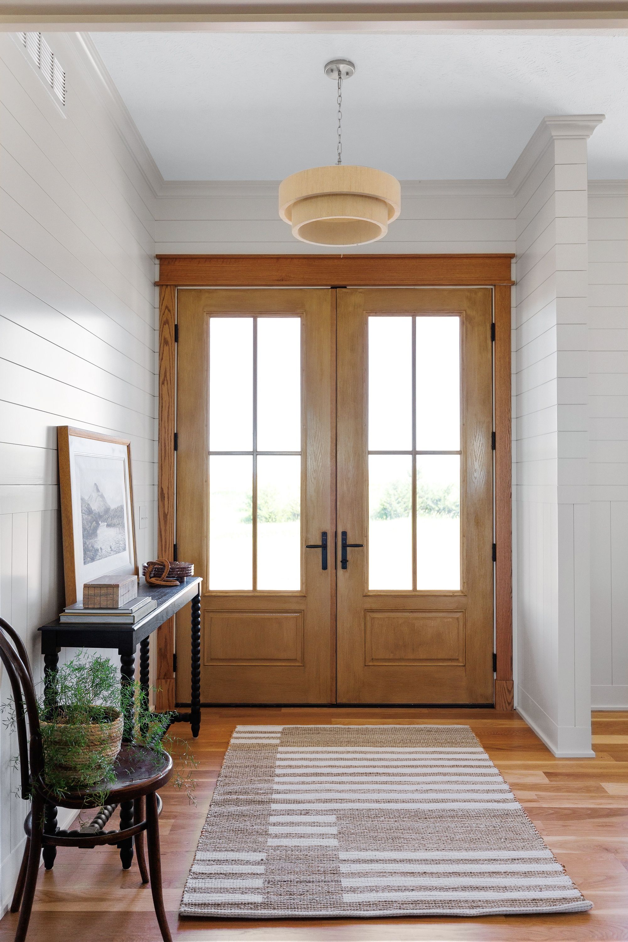 Bright entry with double wood doors flanked by white plank walls, a dark wood console table with a framed picture, and a woven striped rug. A unique tiered light fixture hangs in the center.