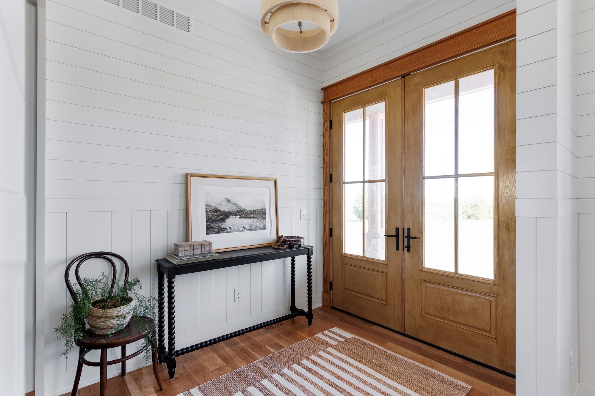 Entryway featuring white shiplap walls, a black console table with a framed landscape painting, and a vintage wooden chair with a potted plant. Double wooden doors let in natural light, and a striped rug adds texture to the hardwood floor.