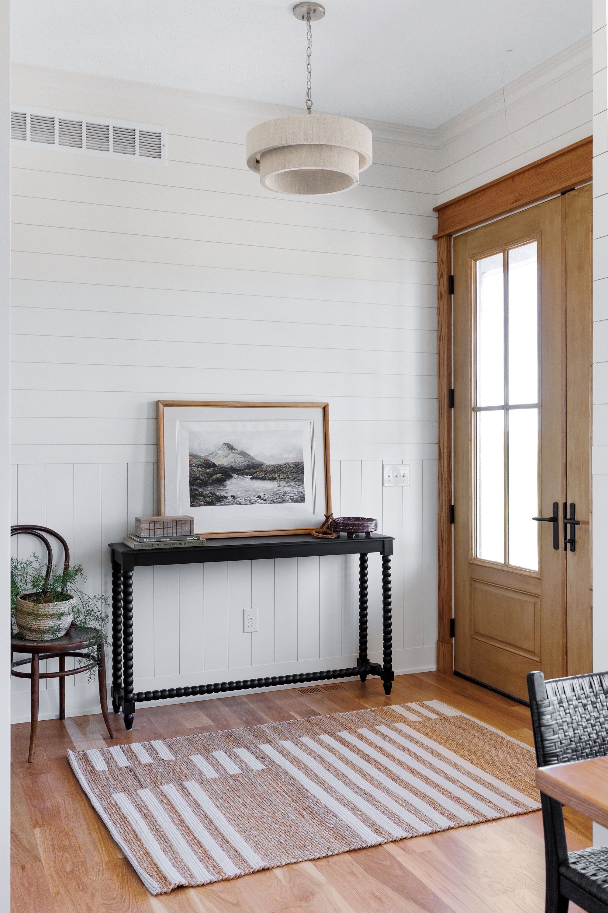 Entryway with white shiplap walls, a black console table with a framed landscape print, and a striped rug. A light wood door with a glass panel is to the right, and a modern pendant light hangs from the ceiling.