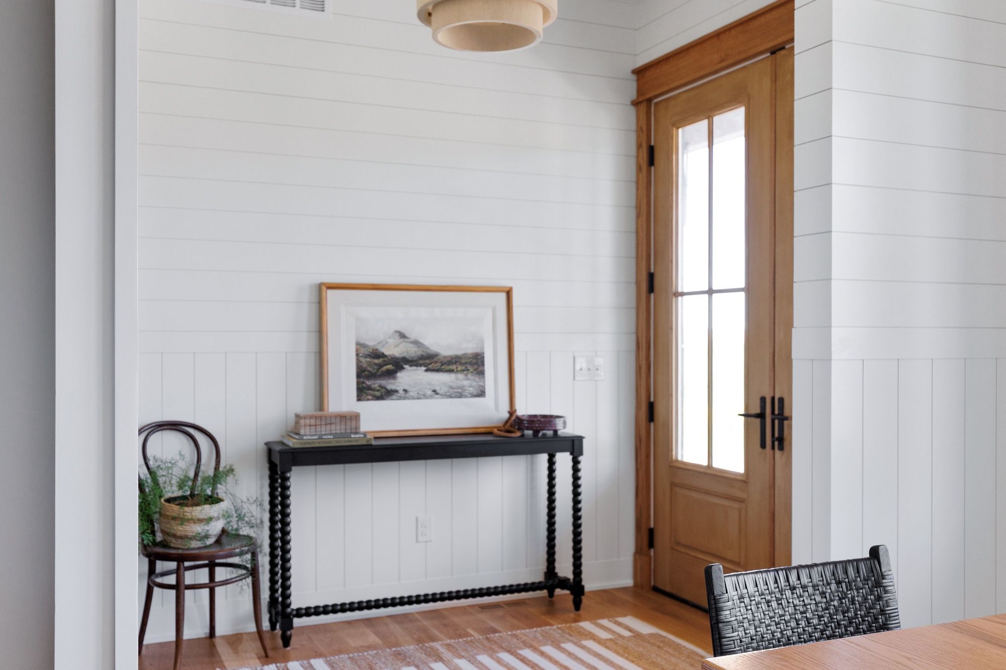 Entryway featuring a black console table with turned legs, topped with a framed landscape print and decorative objects. A small wooden chair holding a potted plant sits nearby, complemented by white shiplap walls and a natural wood door.