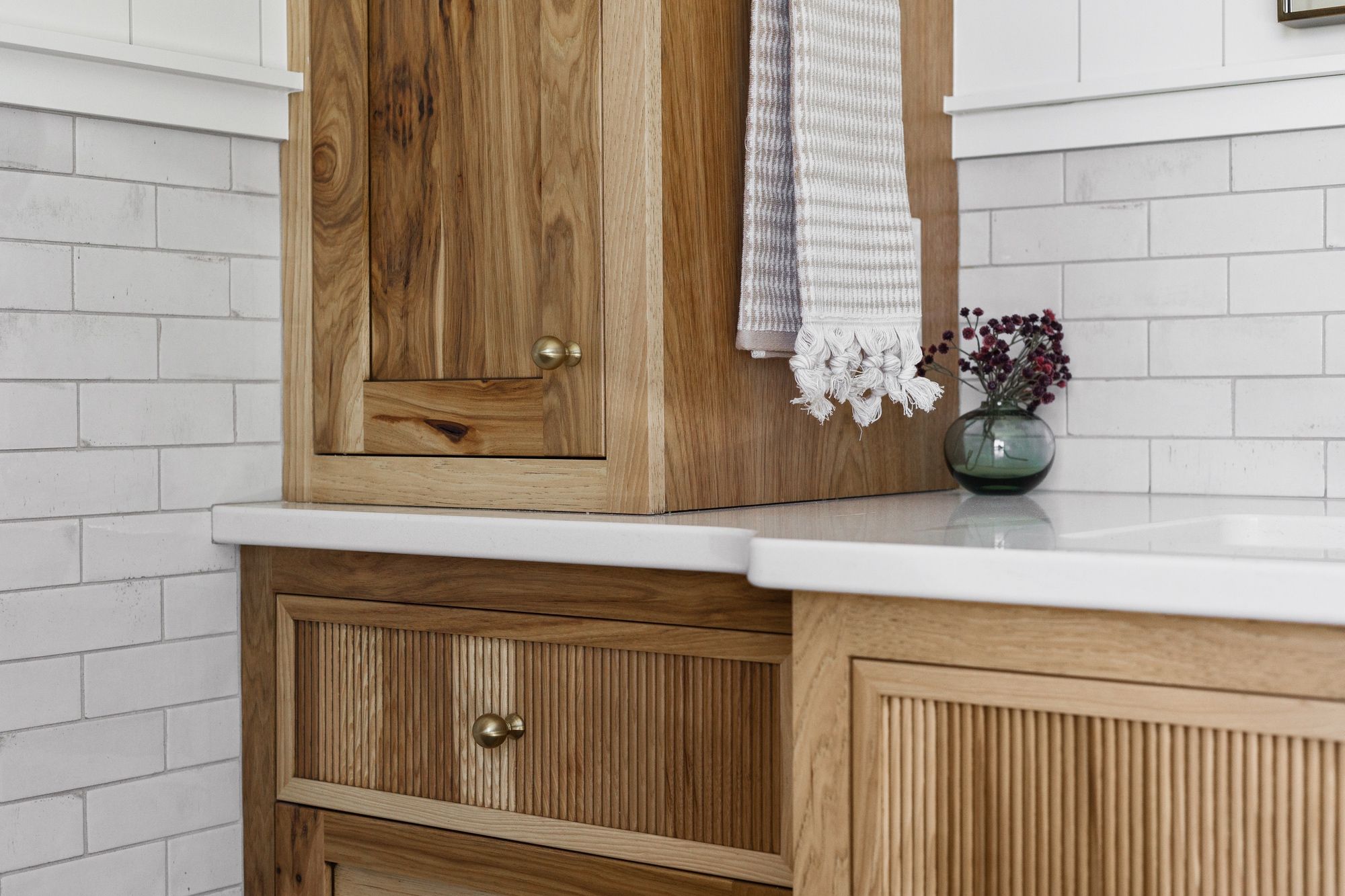 Close-up of a kitchen area featuring light wood cabinets with fluted drawer fronts and brass hardware. A white quartz countertop complements the white subway tile backsplash.